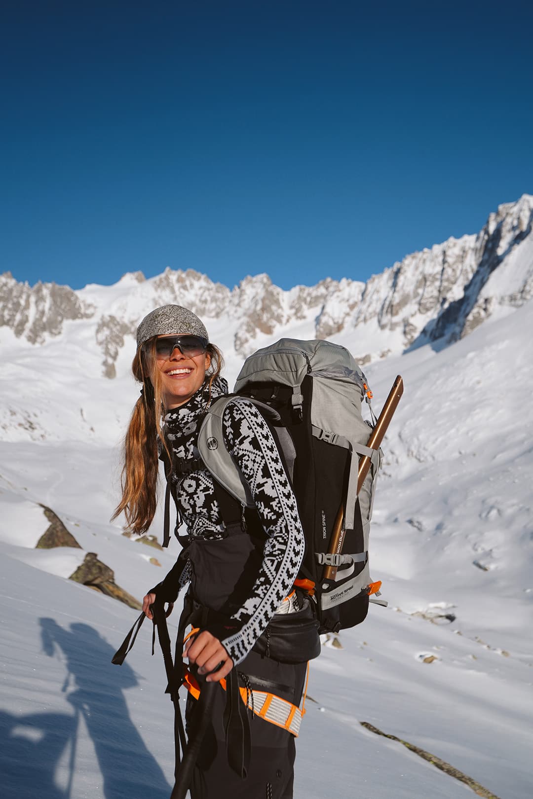 Woman in winter gear smiles while hiking in snowy mountains, carrying a large backpack and using trekking poles under a clear blue sky.