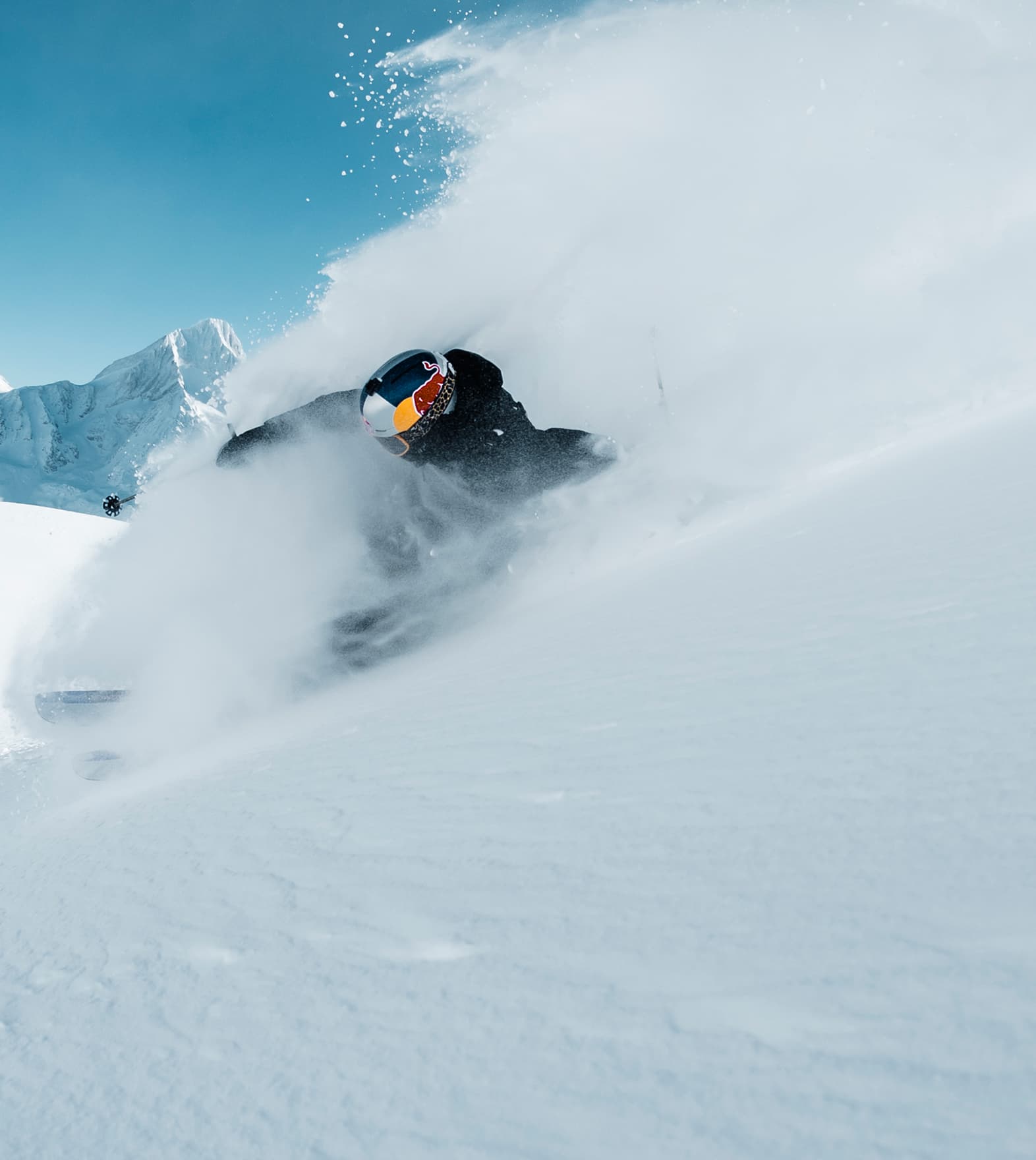 A skier in black gear and a reflective visor goggles is carving through fresh powder snow on a sunny, mountainous slope.