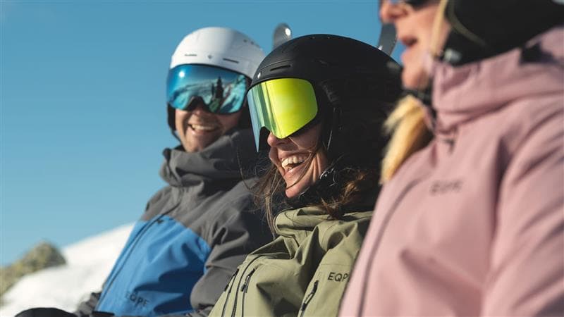 Three people in winter gear and helmets smile while sitting on a ski lift, with a clear blue sky in the background.