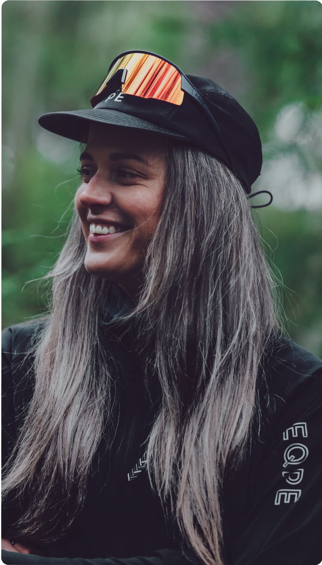 Smiling woman with long hair, wearing a black cap and sunglasses.