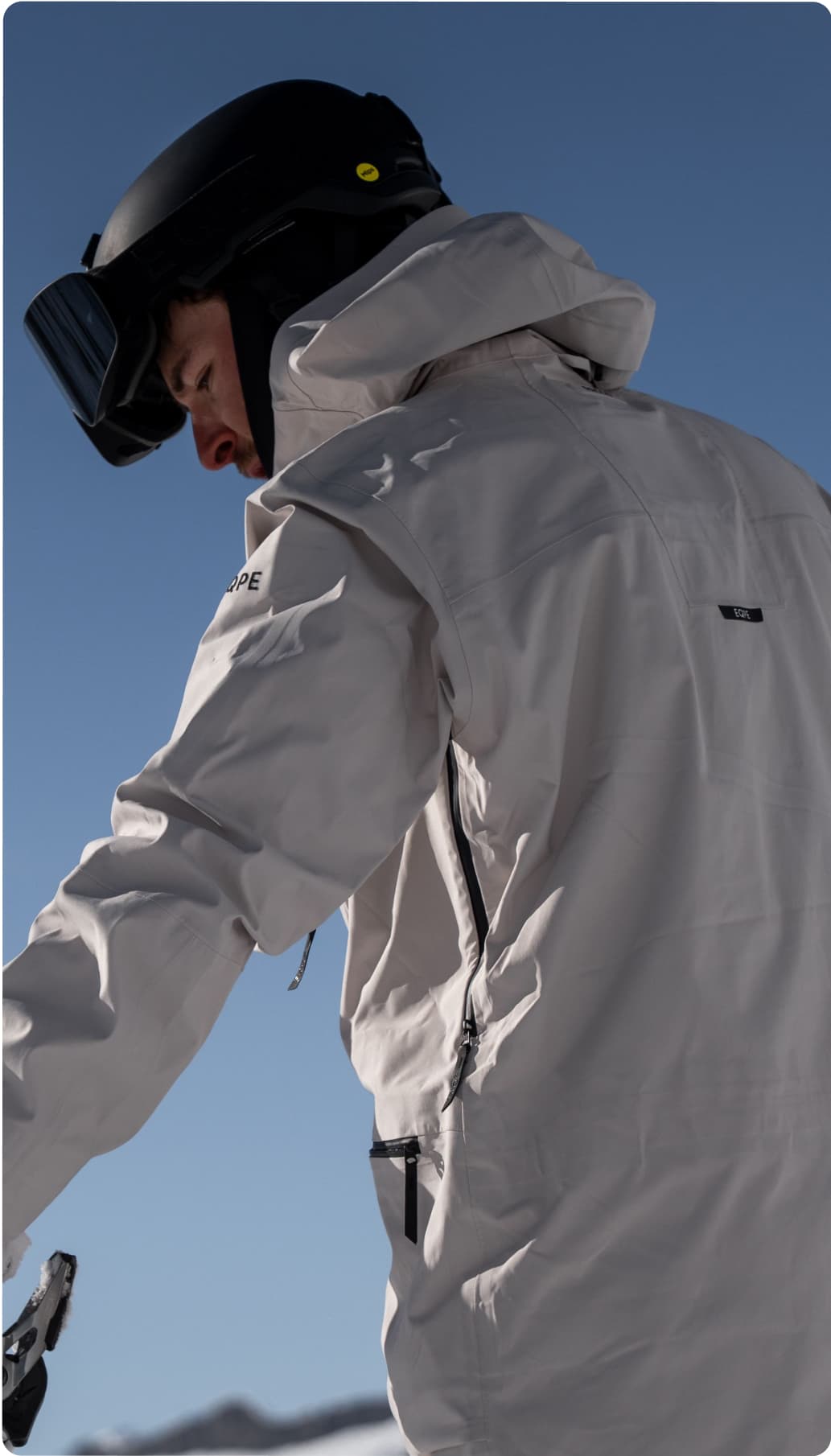 Skiier in a white beige ski jacket and black helmet stands against a clear blue sky.