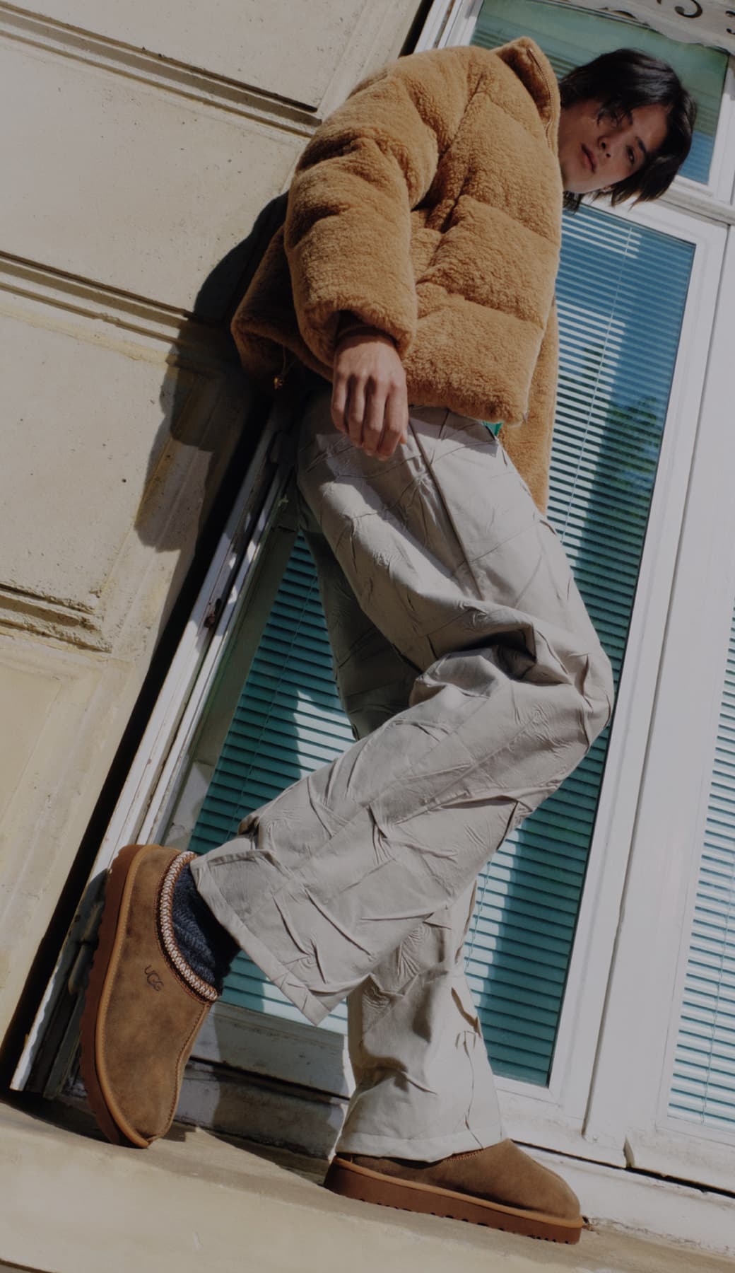 Person in a brown fleece jacket and beige pants leans against a building.