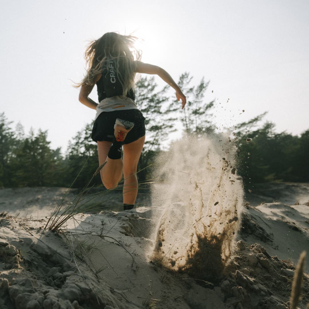 Woman running on a sandy beach, kicking up sand, with trees and a bright sky in the background.