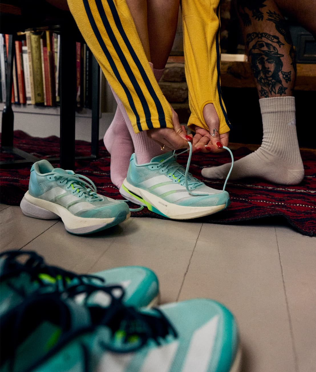 Person in yellow jacket tying laces on teal sneakers, sitting beside another person with tattoos and socks, on a patterned rug.