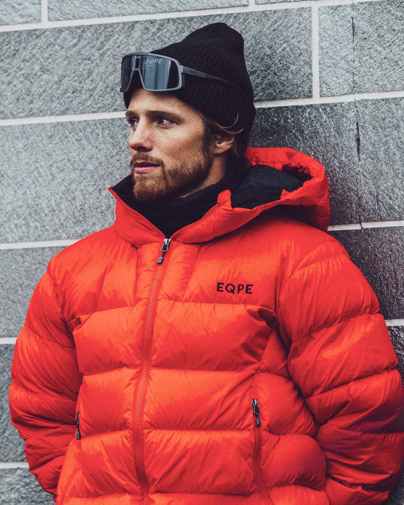 Man in a red puffer jacket and black beanie stands against a gray brick wall.