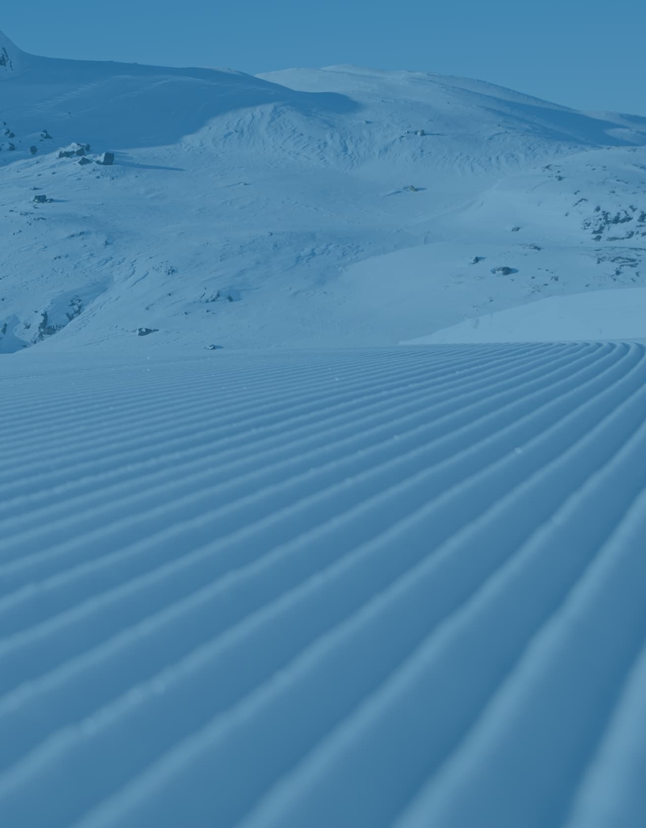 Snow-covered landscape with neatly groomed, parallel ridges in the foreground, leading to distant snow-covered hills under a clear blue sky.