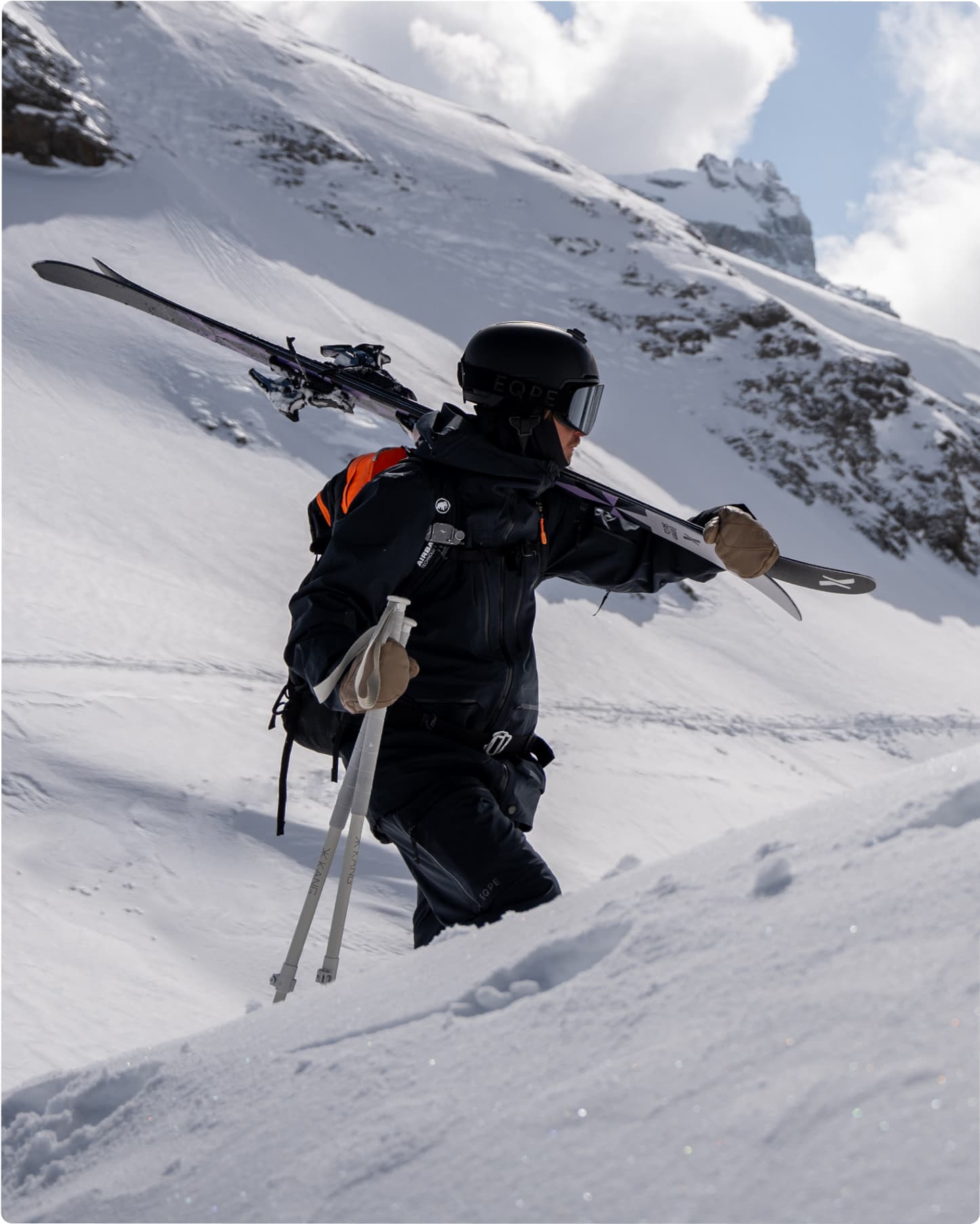 A skier in black gear climbs a snowy mountain with skis on their shoulder.