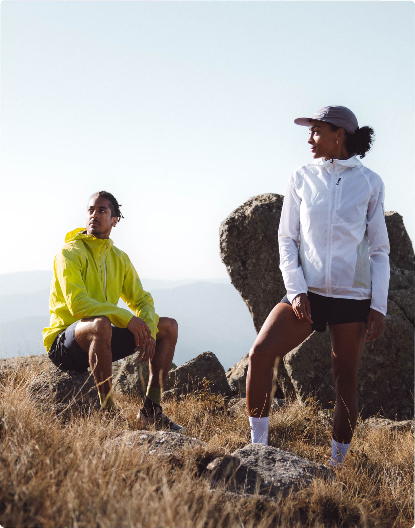 Two people in athletic outfits rest on a mountaintop, overlooking a vast landscape.