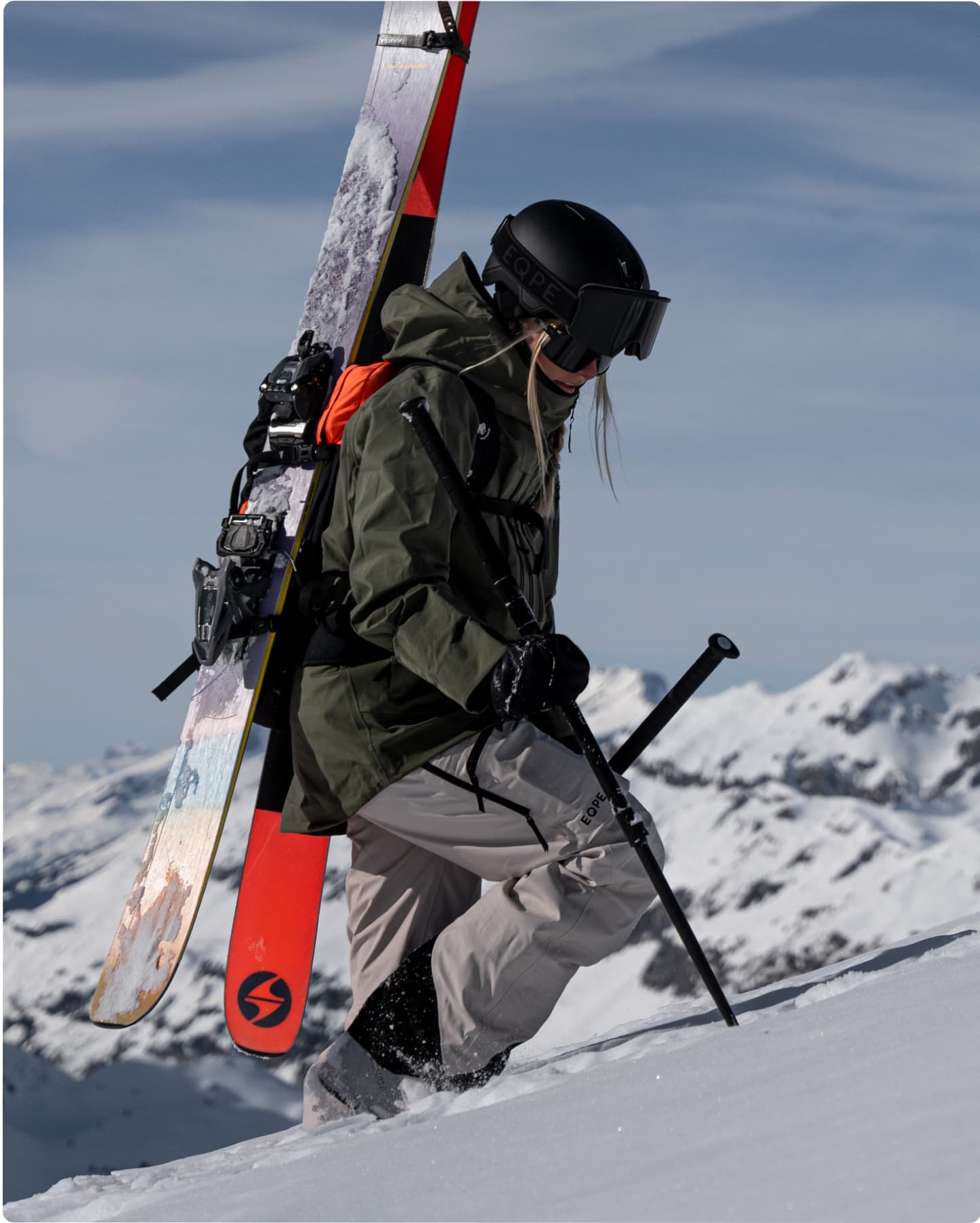 Skier hiking up a snowy mountain wearing a green ski jacket and grey ski pants.