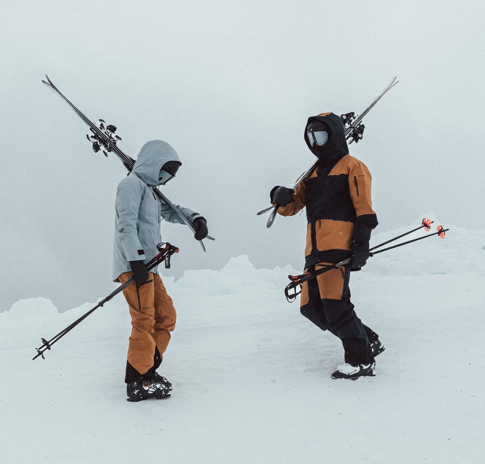 Two people in ski gear stand on a snowy mountain slope under a cloudy sky.