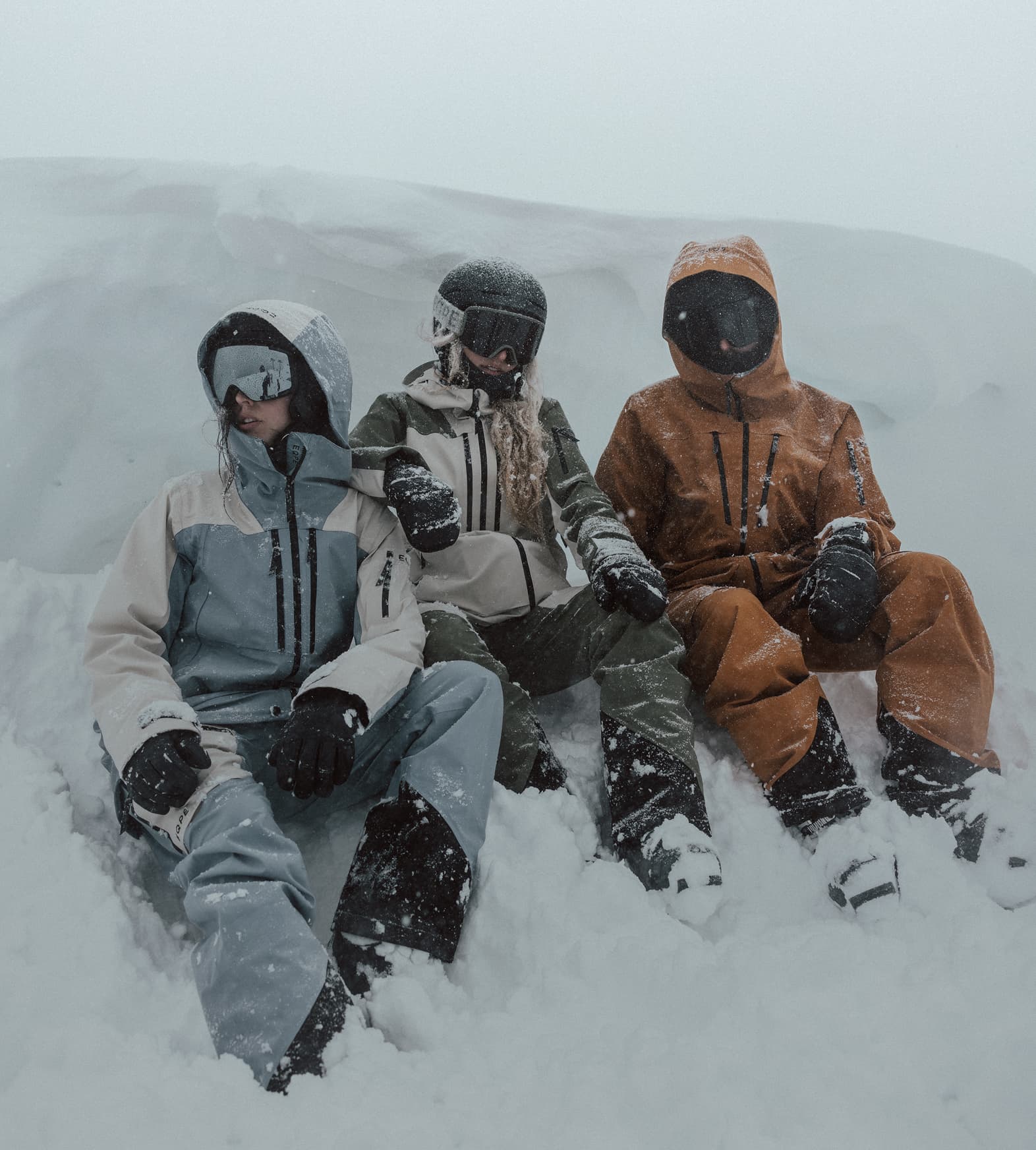Three people in ski wear, sit in the snow during a snowfall.