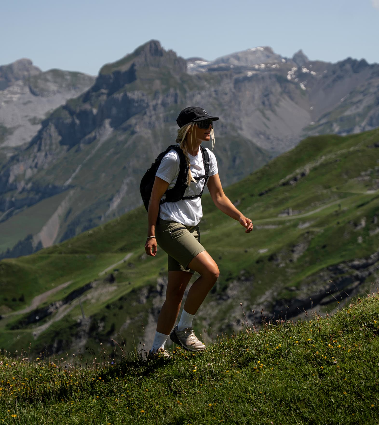 Woman hiking on a grassy mountain trail, with rugged peaks in the background.