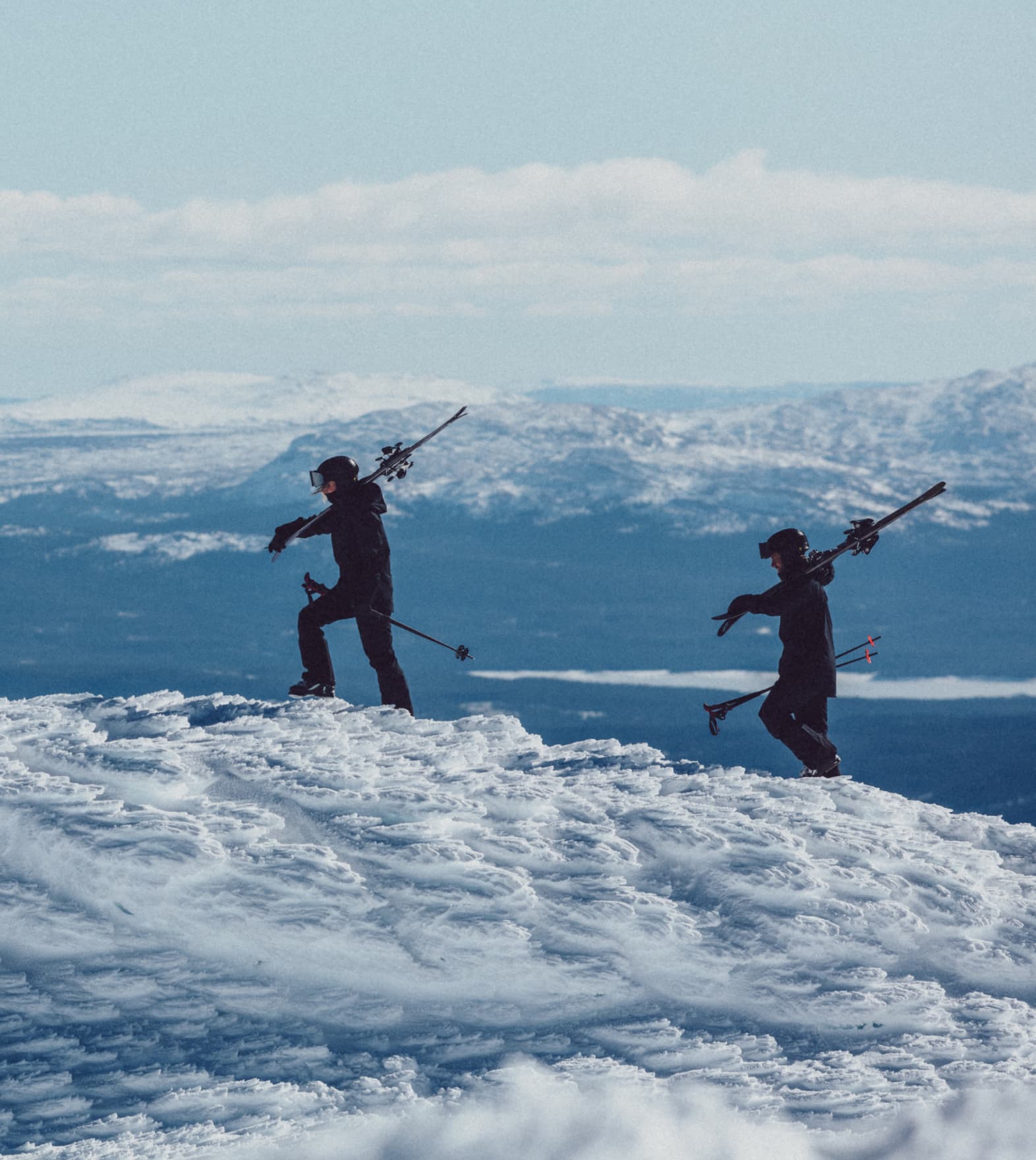 Two skiers in dark ski gear carry skis on their shoulders, hiking up a snowy hill with a mountain range in the background.