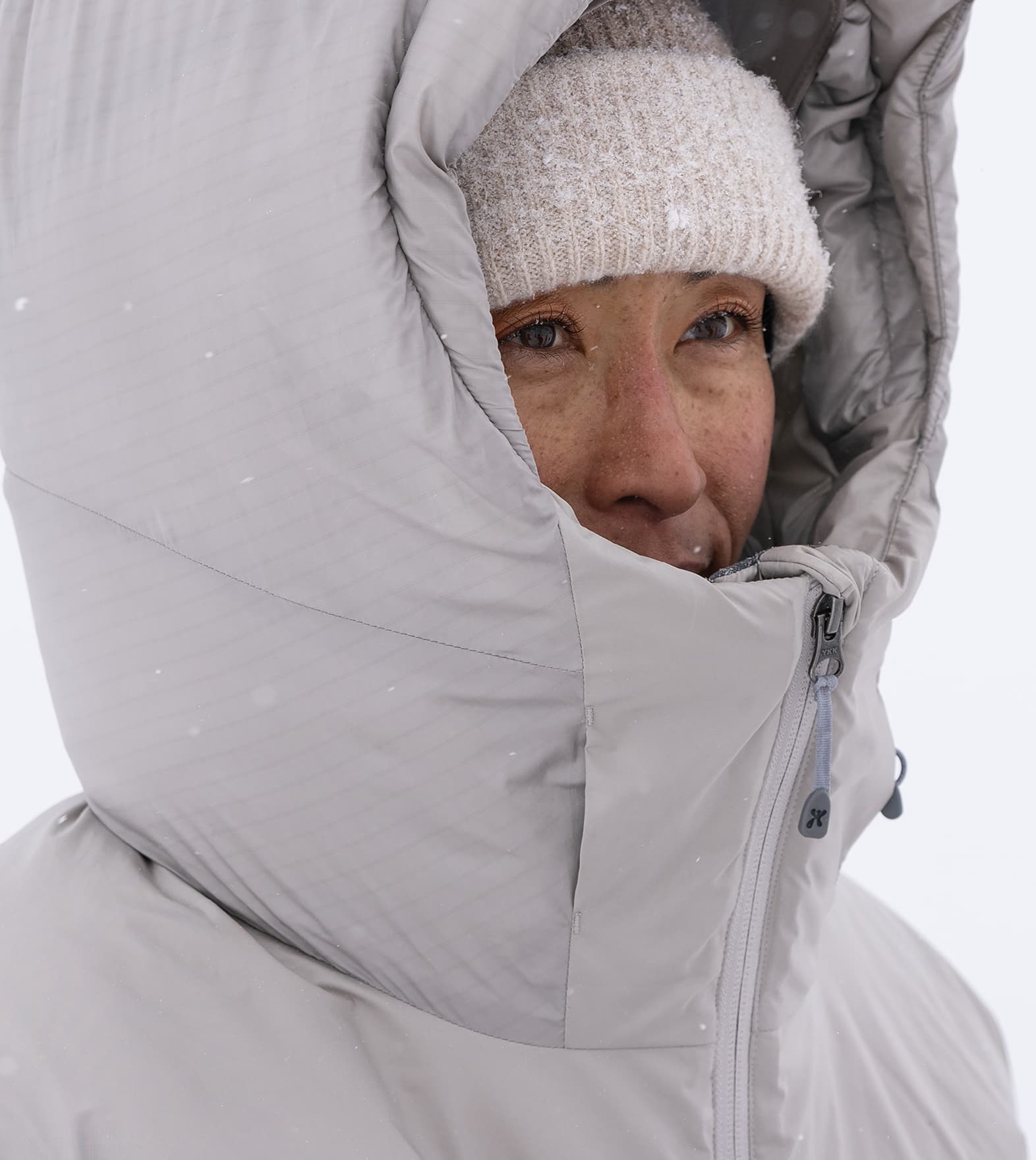 Person bundled in a thick, gray winter coat and white knit hat, with snowflakes falling, against a snowy background.