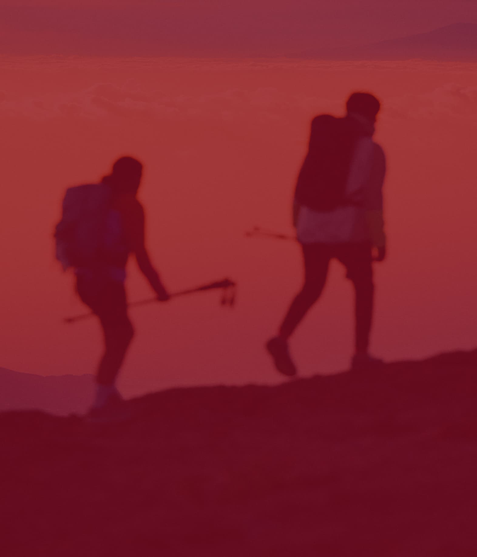Two silhouettes of hikers with backpacks and trekking poles walking along a mountain ridge.