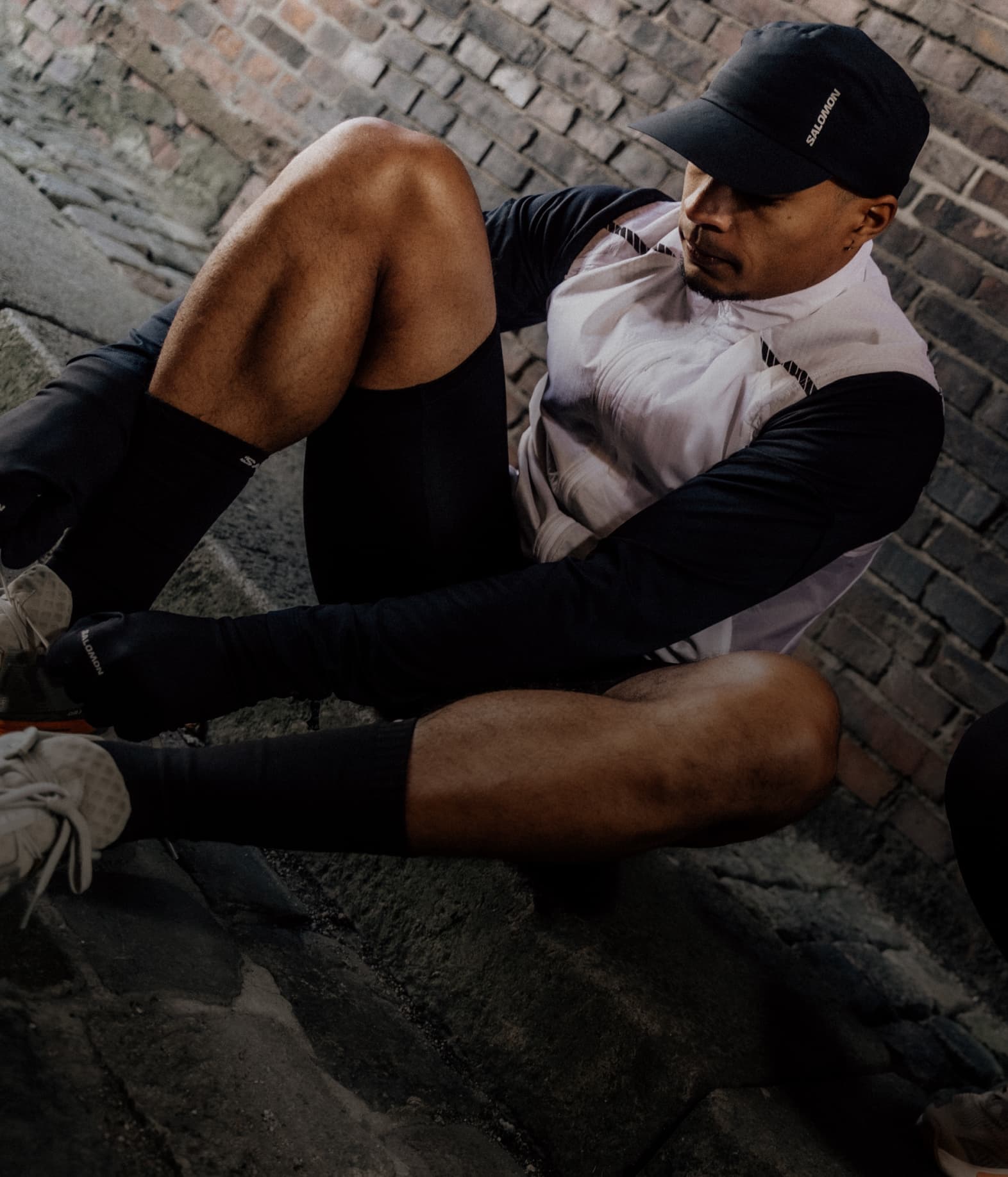 A person in athletic wear, sitting on a curb, tying shoelaces, brick walls in the background..