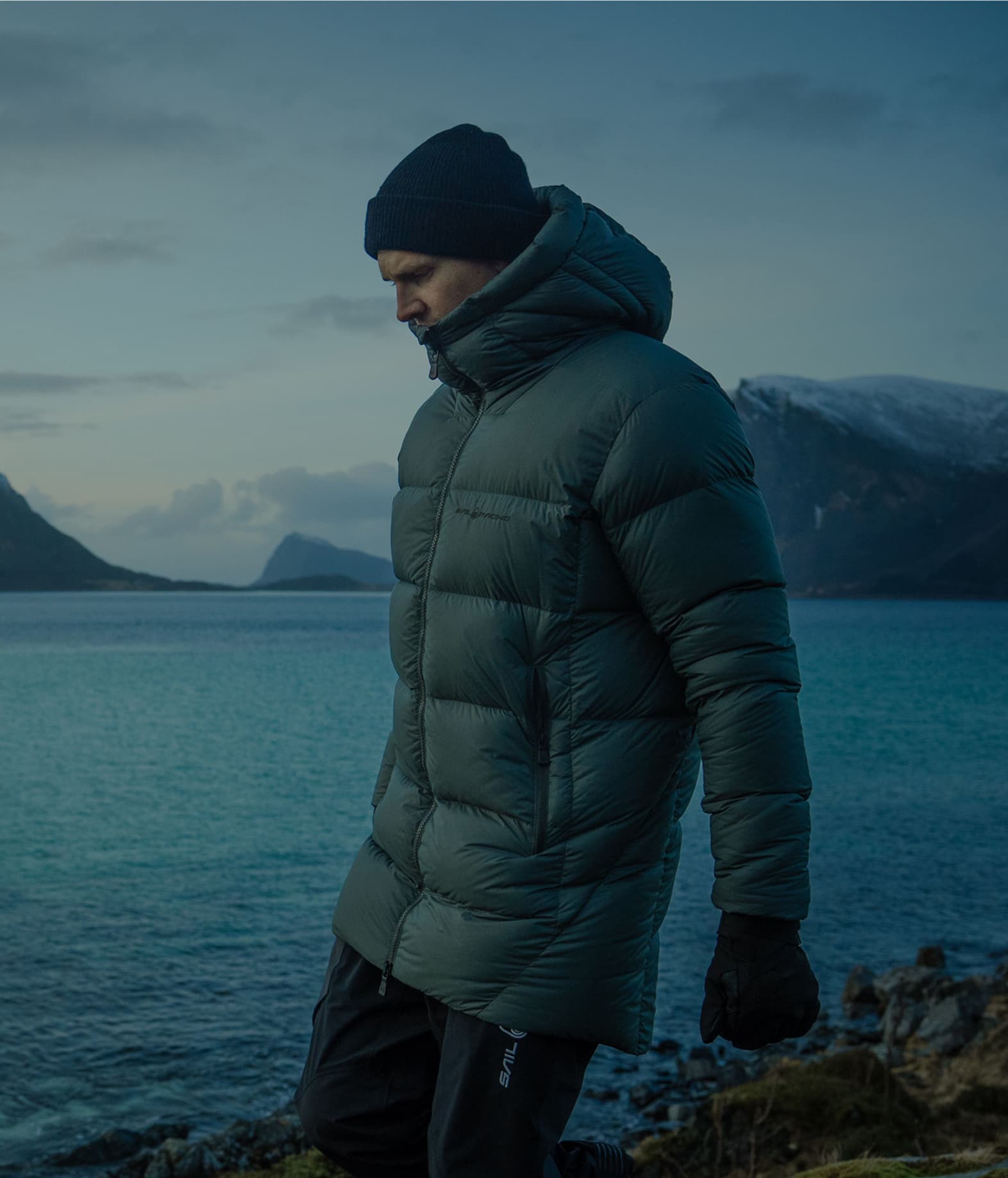 Person in a green puffer jacket and beanie walking by a calm, scenic lake with mountains in the background, under a cloudy sky.