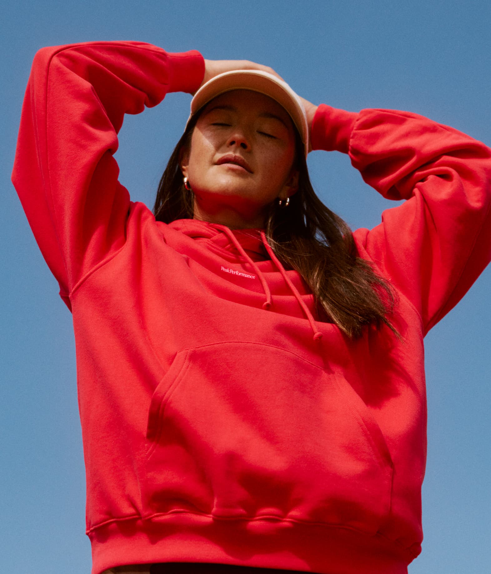 Person in a red hoodie and beige cap standing against a clear blue sky, with eyes closed and hands resting on their head.