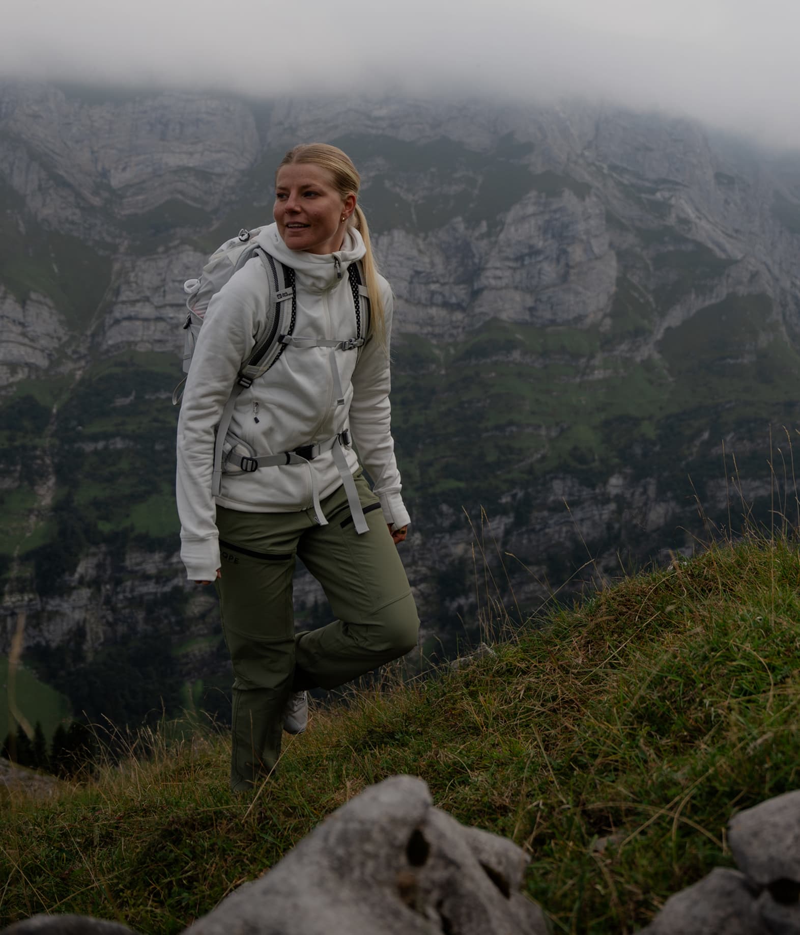 A person in hiking gear stands on a grassy mountainside, with misty mountains in the background.