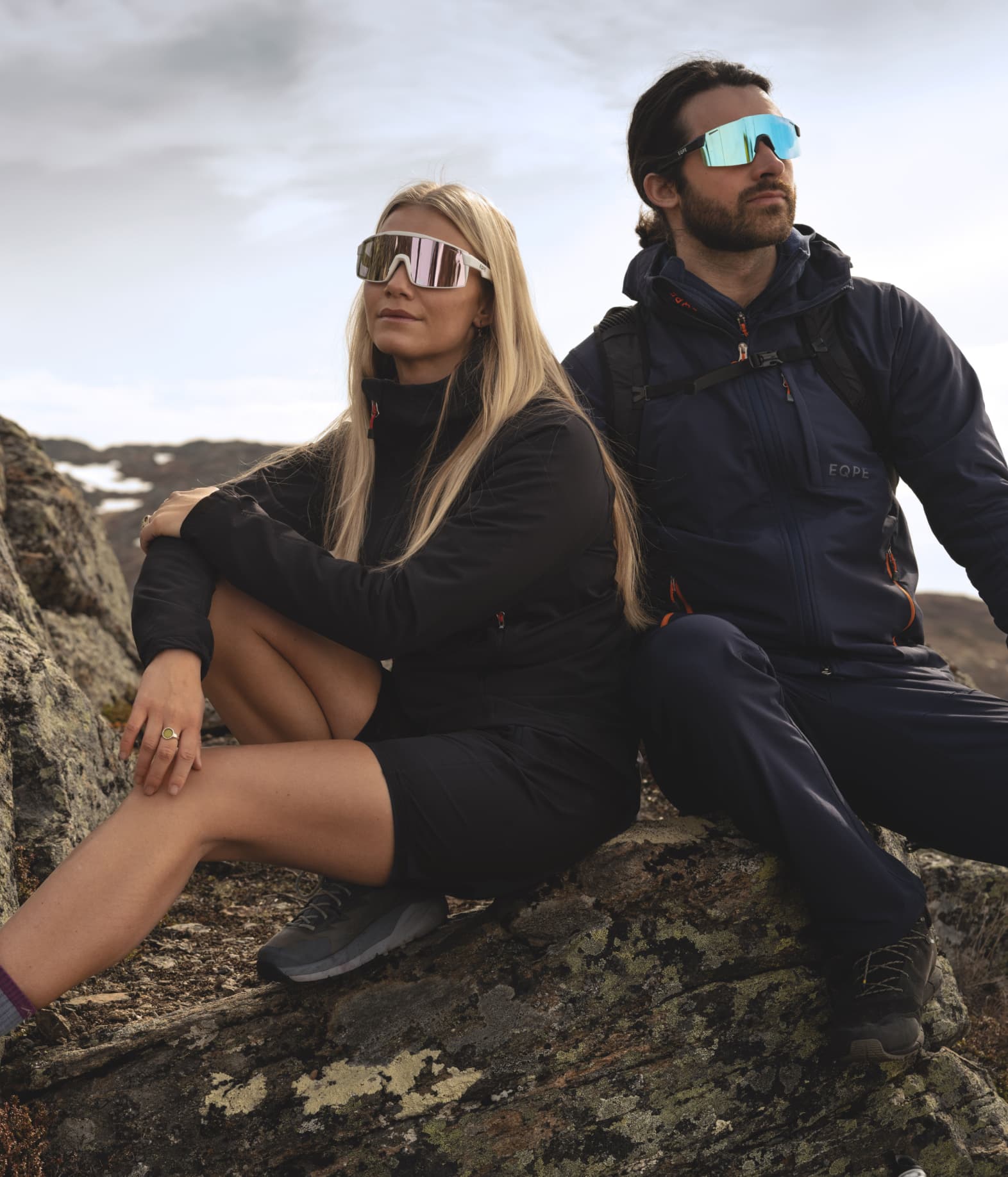 Two people in outdoor gear sit on rocky terrain, gazing into the distance.