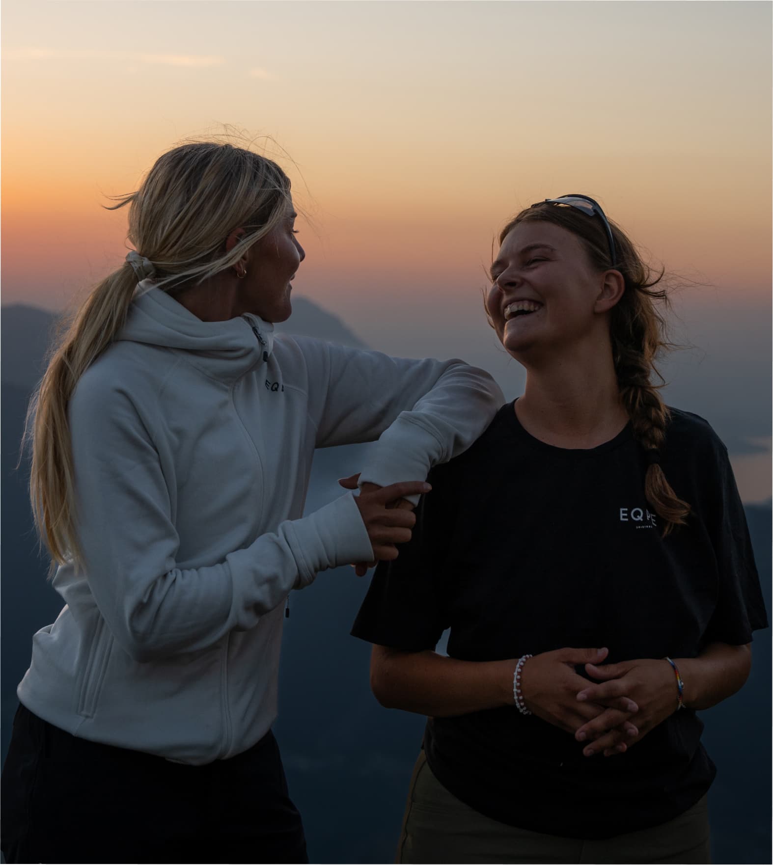 Two people smiling at each other, standing on a mountain at sunset with a view of distant hills and a glowing sky.