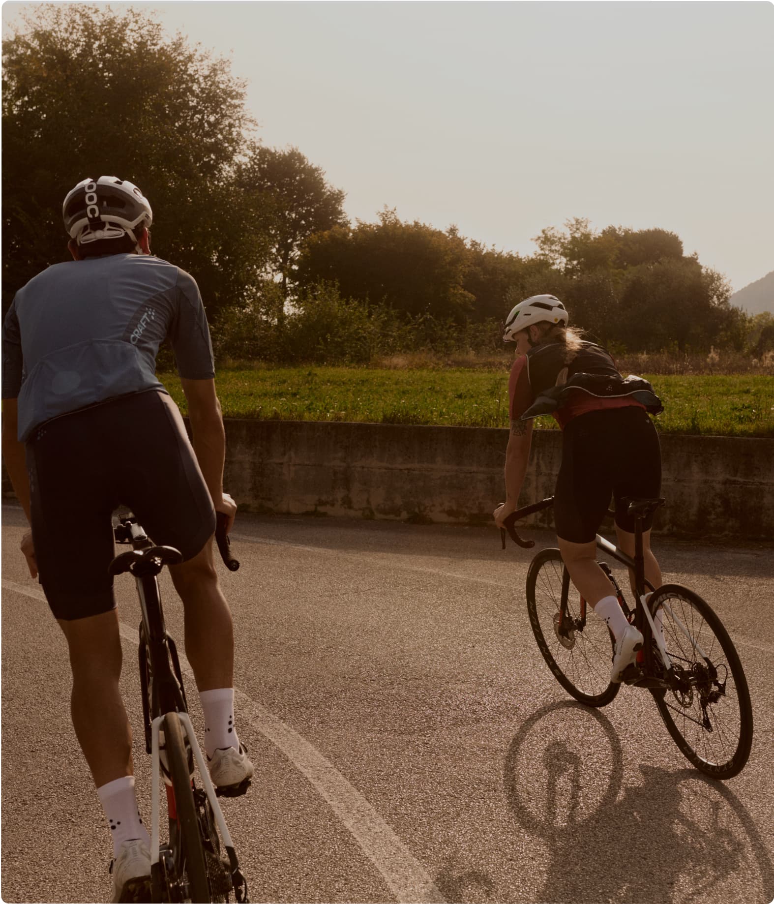 Two cyclists ride on a road beside a grassy field with trees.
