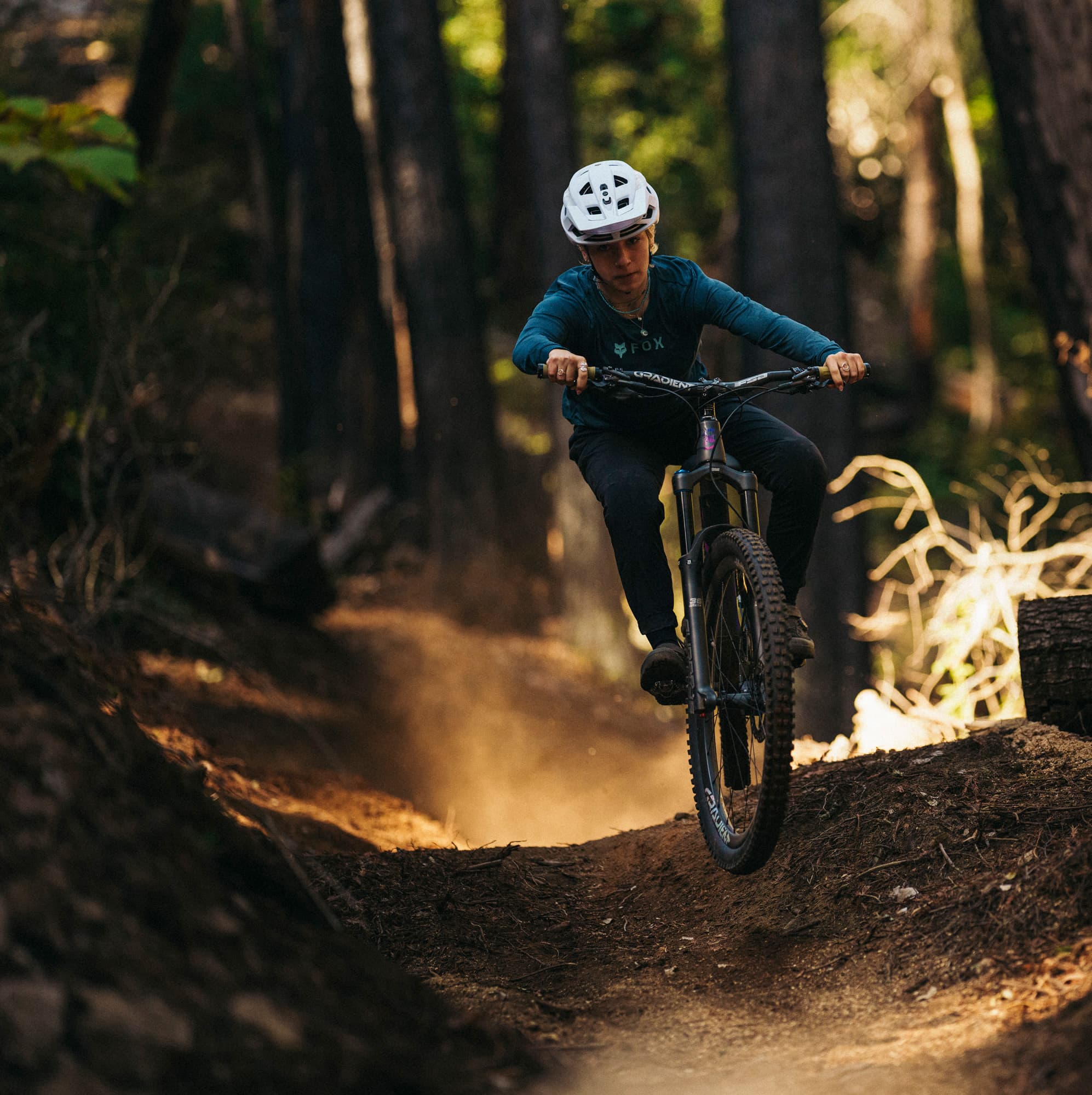 Person mountain biking on a forest trail, wearing a white helmet and blue shirt, with sunlight filtering through the trees.