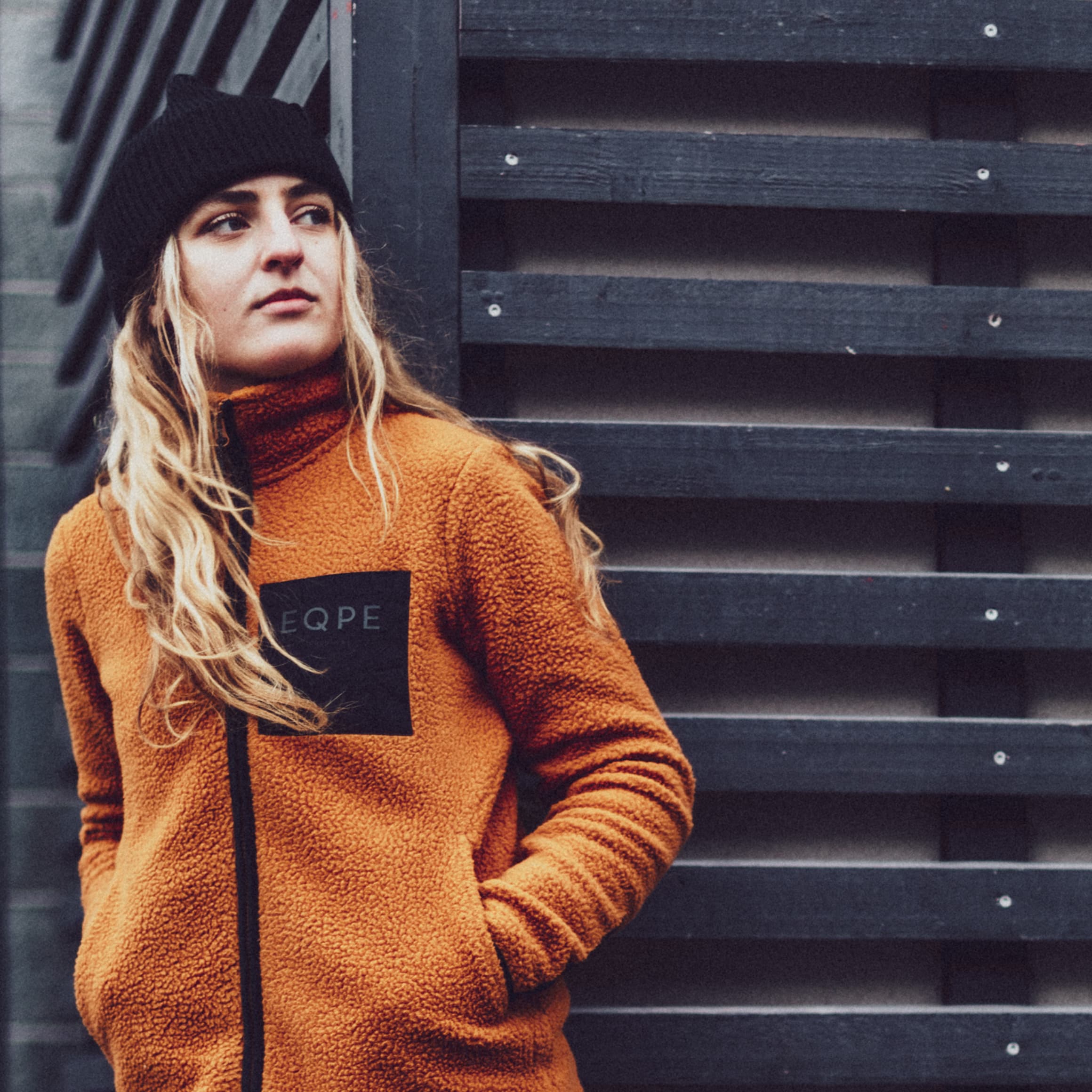 Woman in an orange fleece jacket and black beanie stands against a dark wooden wall, looking to the side.