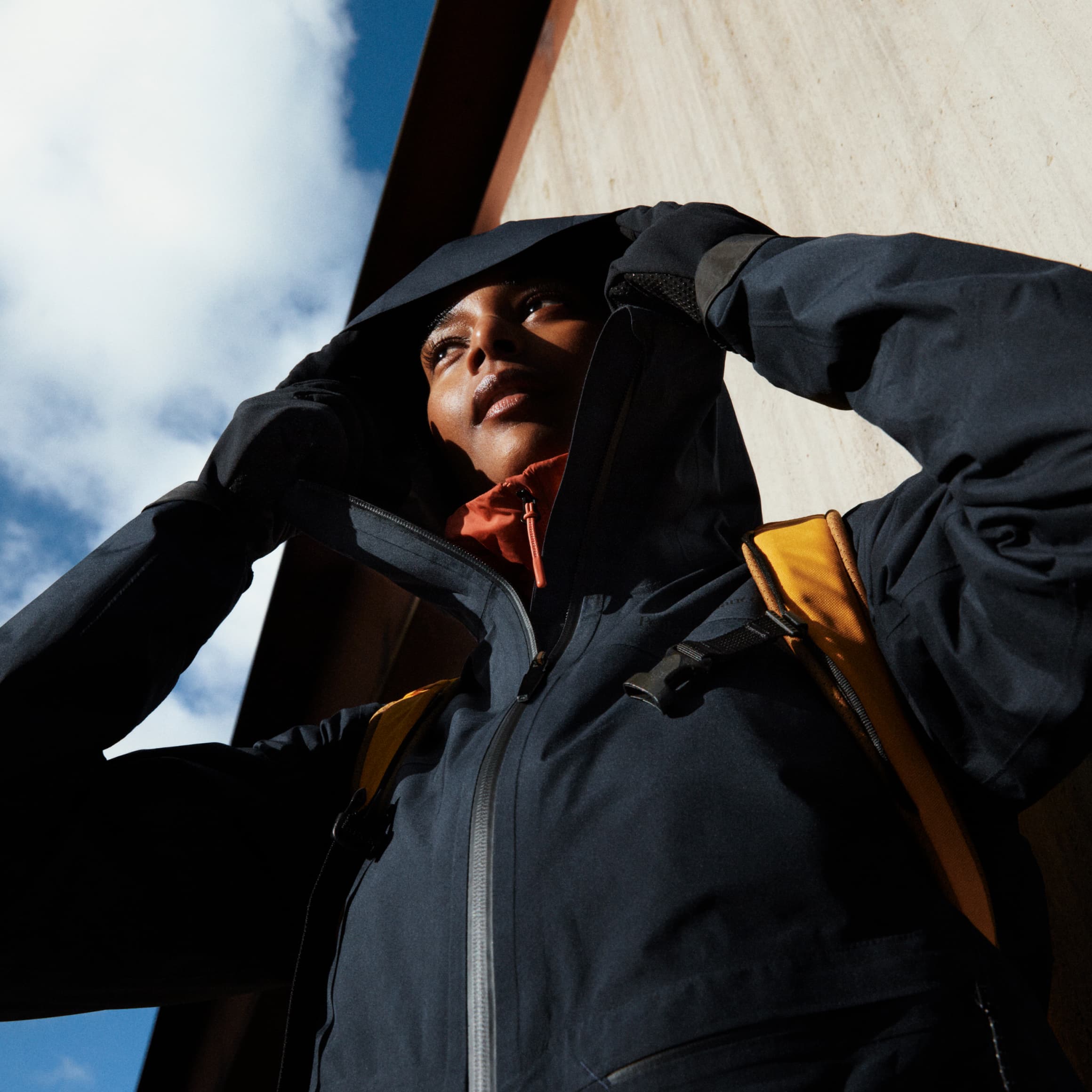 Person in a dark hooded jacket and yellow backpack looks up, standing against a blue sky and building.