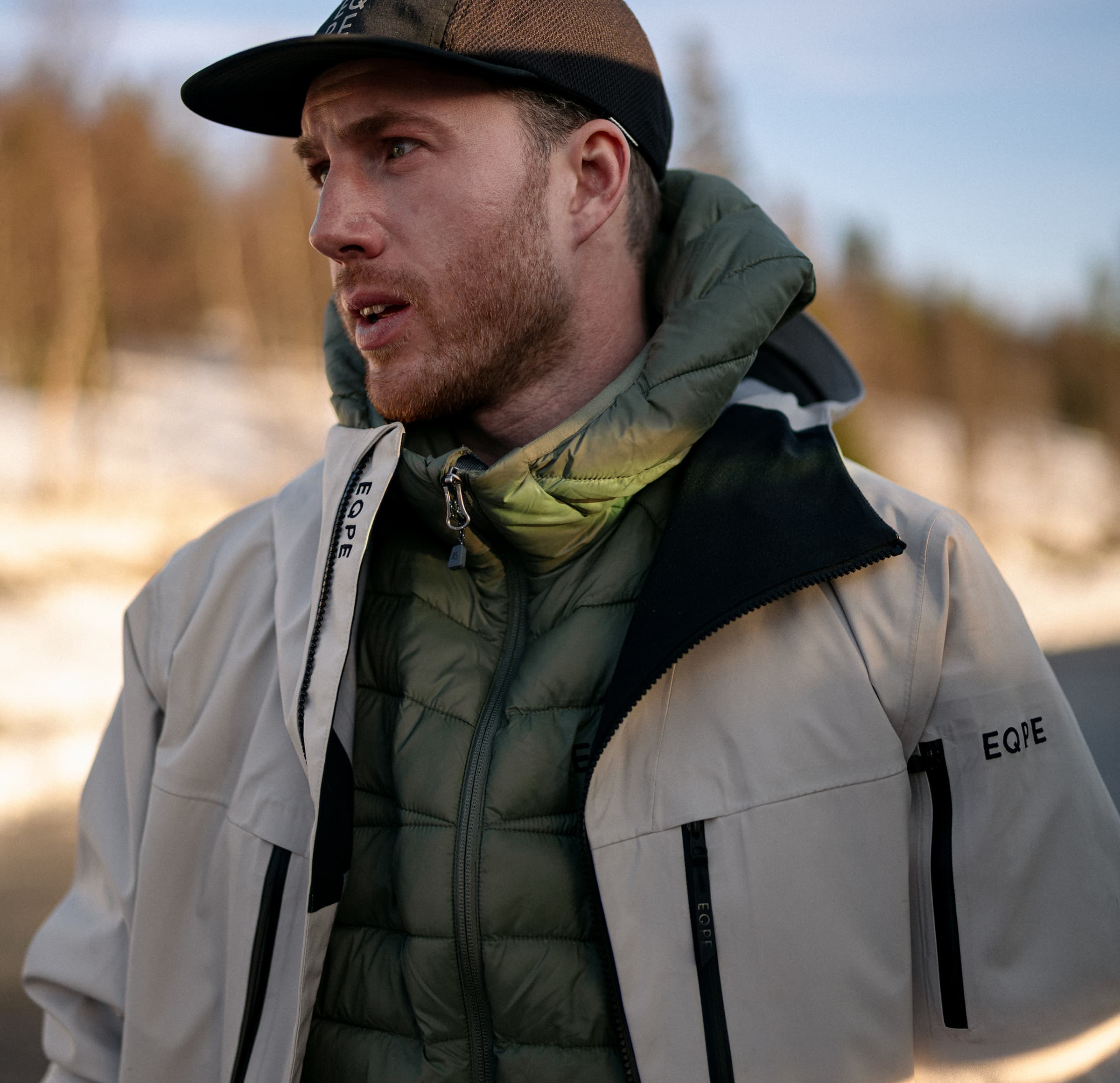 Man in a beige jacket and green puffer, stands outdoors with a blurred snowy background.