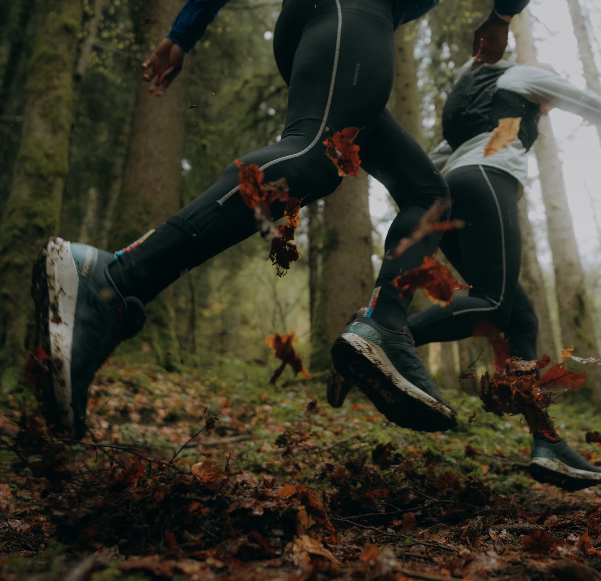 Two people trail running through a forest, kicking up fallen leaves. They wear athletic gear and the scene conveys motion and energy.