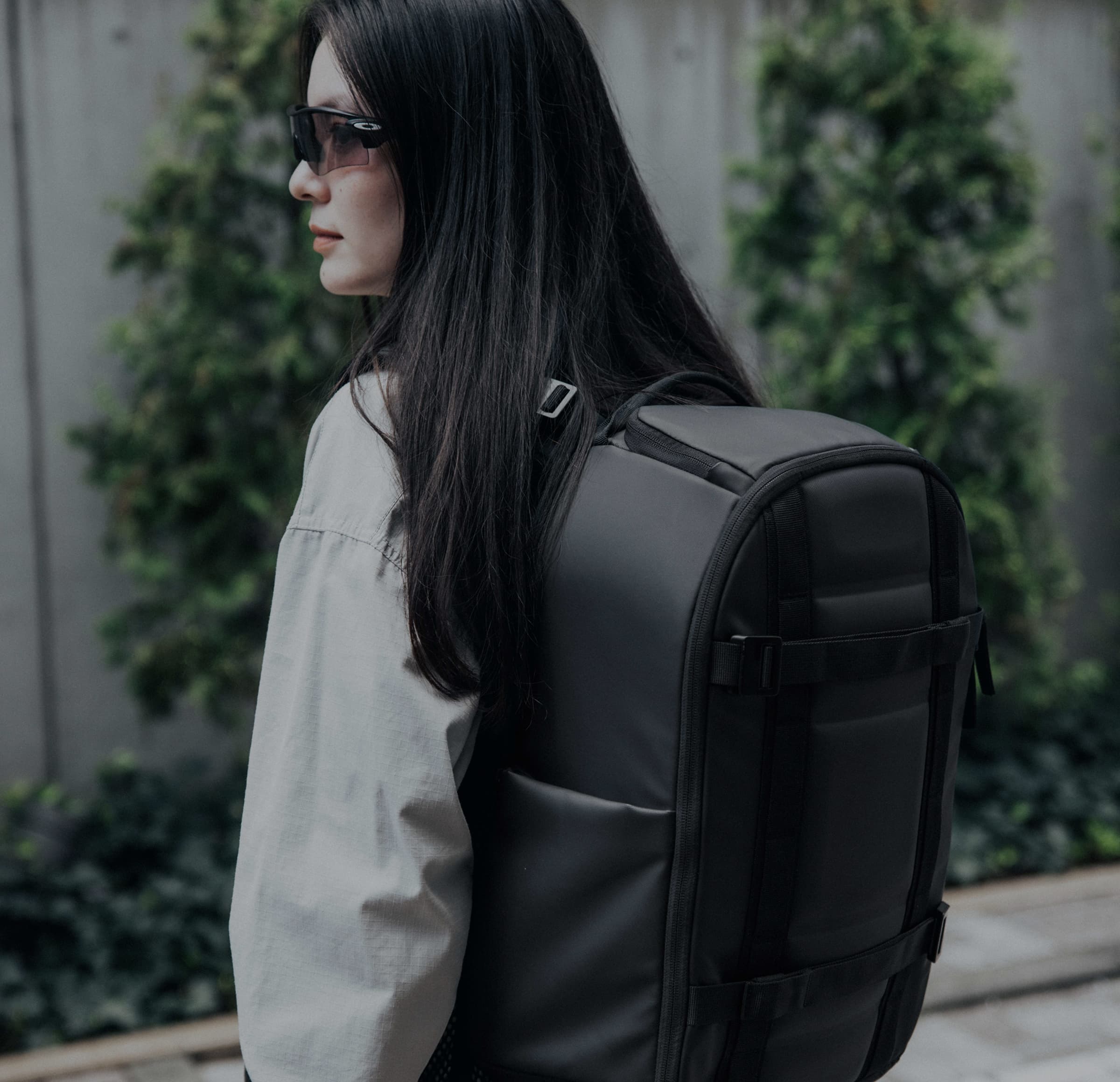 A woman wearing sunglasses and a light jacket carries a large black backpack outdoors, with greenery in the background.