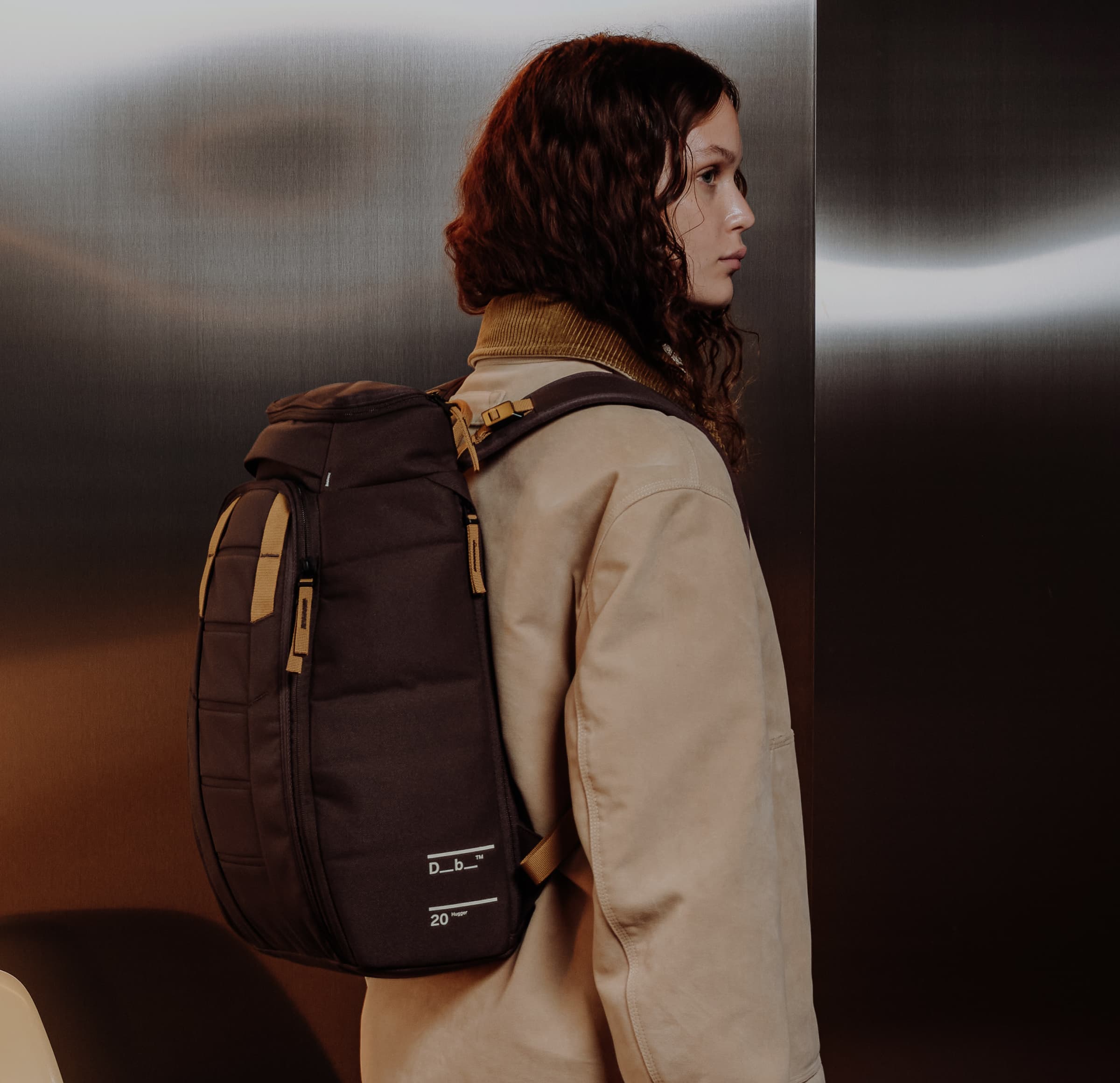 Person wearing a beige coat and carrying a dark backpack, standing in front of a metallic wall.