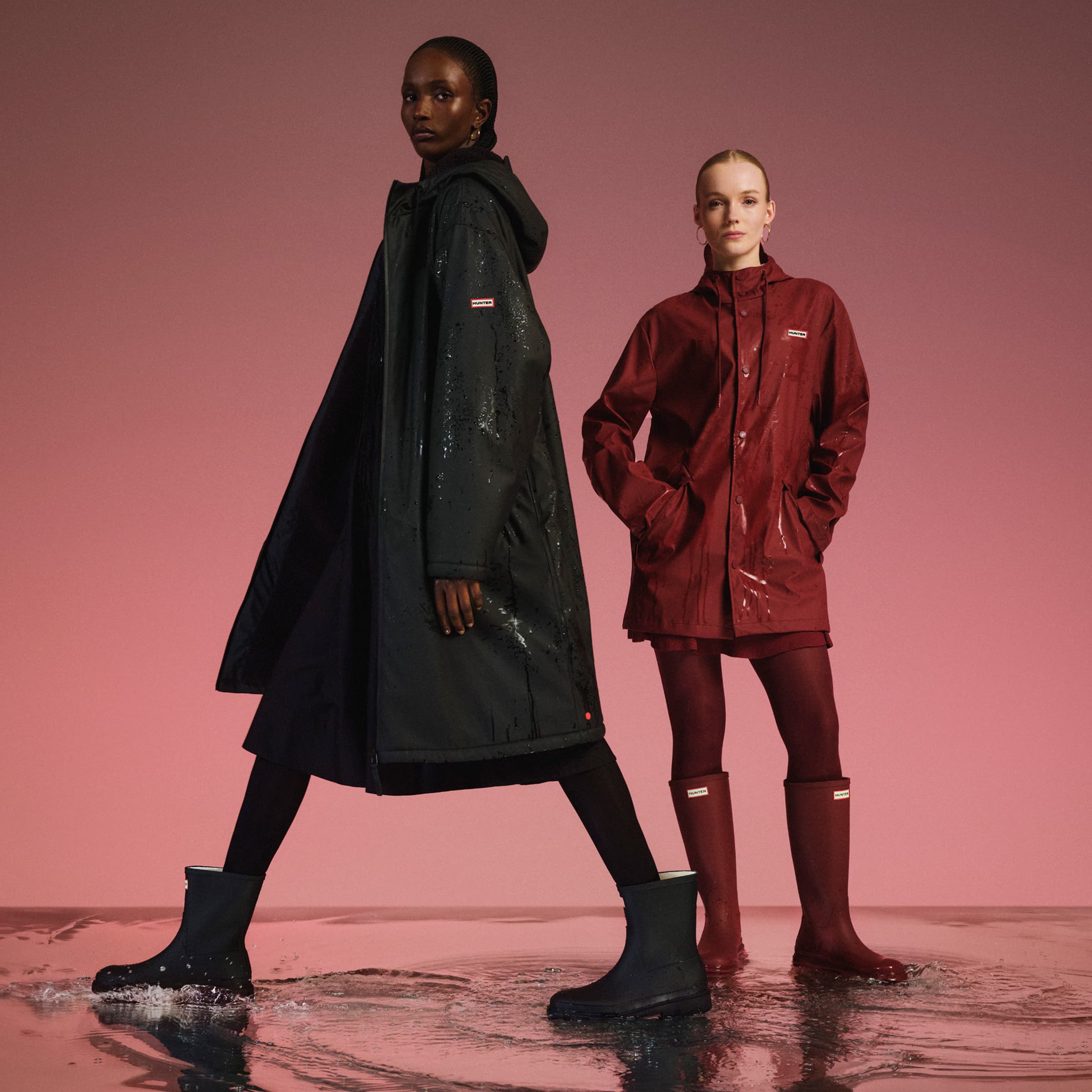 Two models in raincoats and boots stand on a wet surface against a pink background.