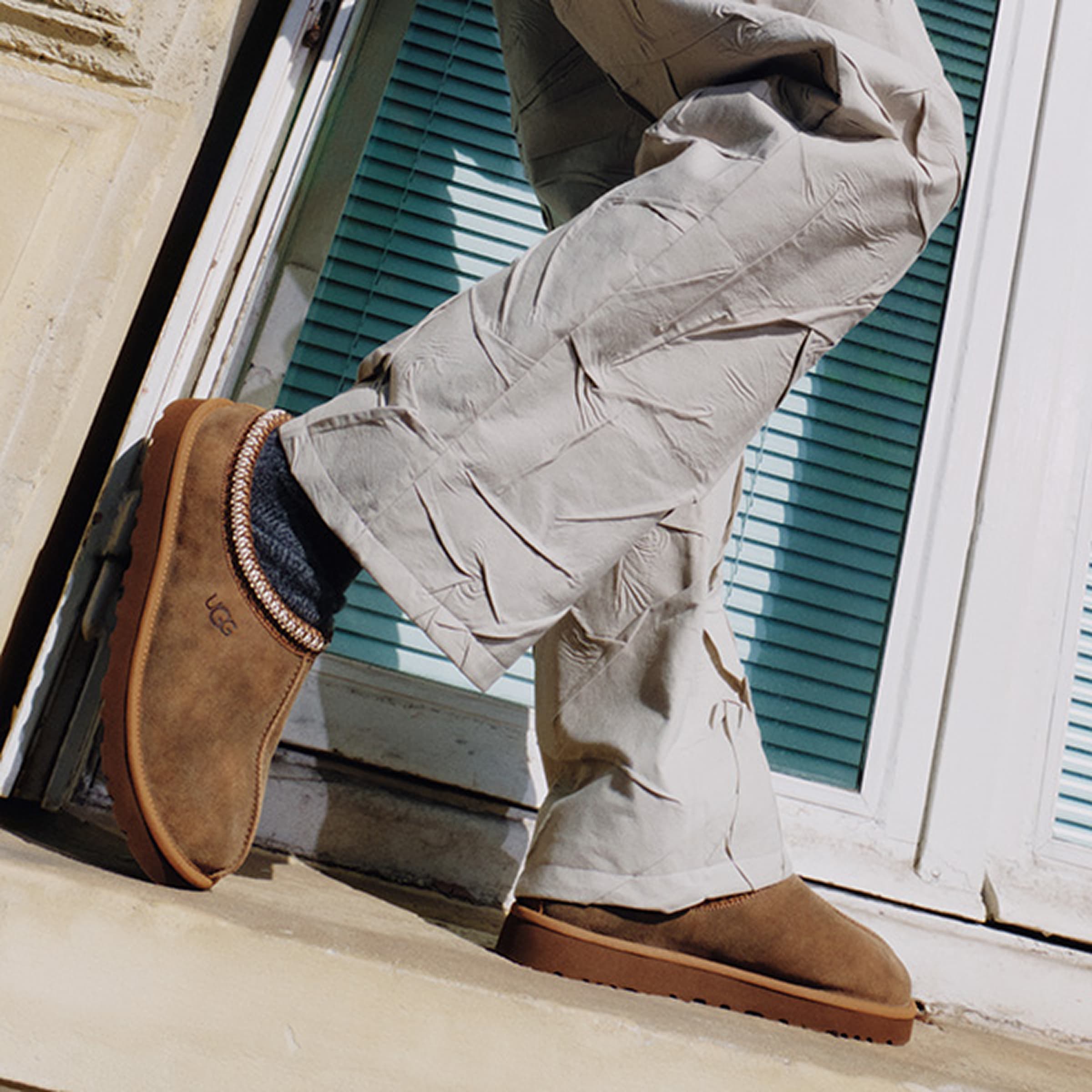 Person wearing beige crinkled pants and brown slip-on shoes, stepping onto a ledge near a window with blue blinds.