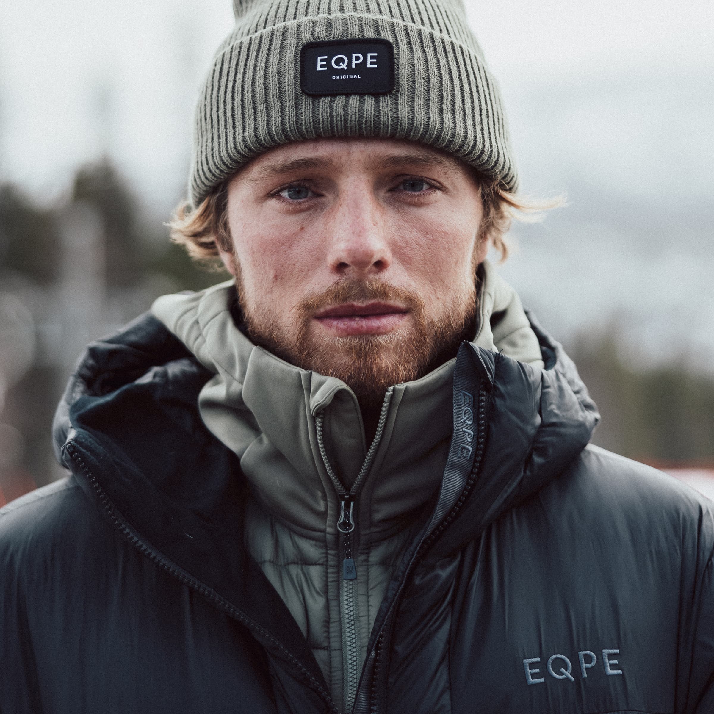 A person wearing a green beanie and a black puffer jacket stands outdoors, with a blurred snowy background.