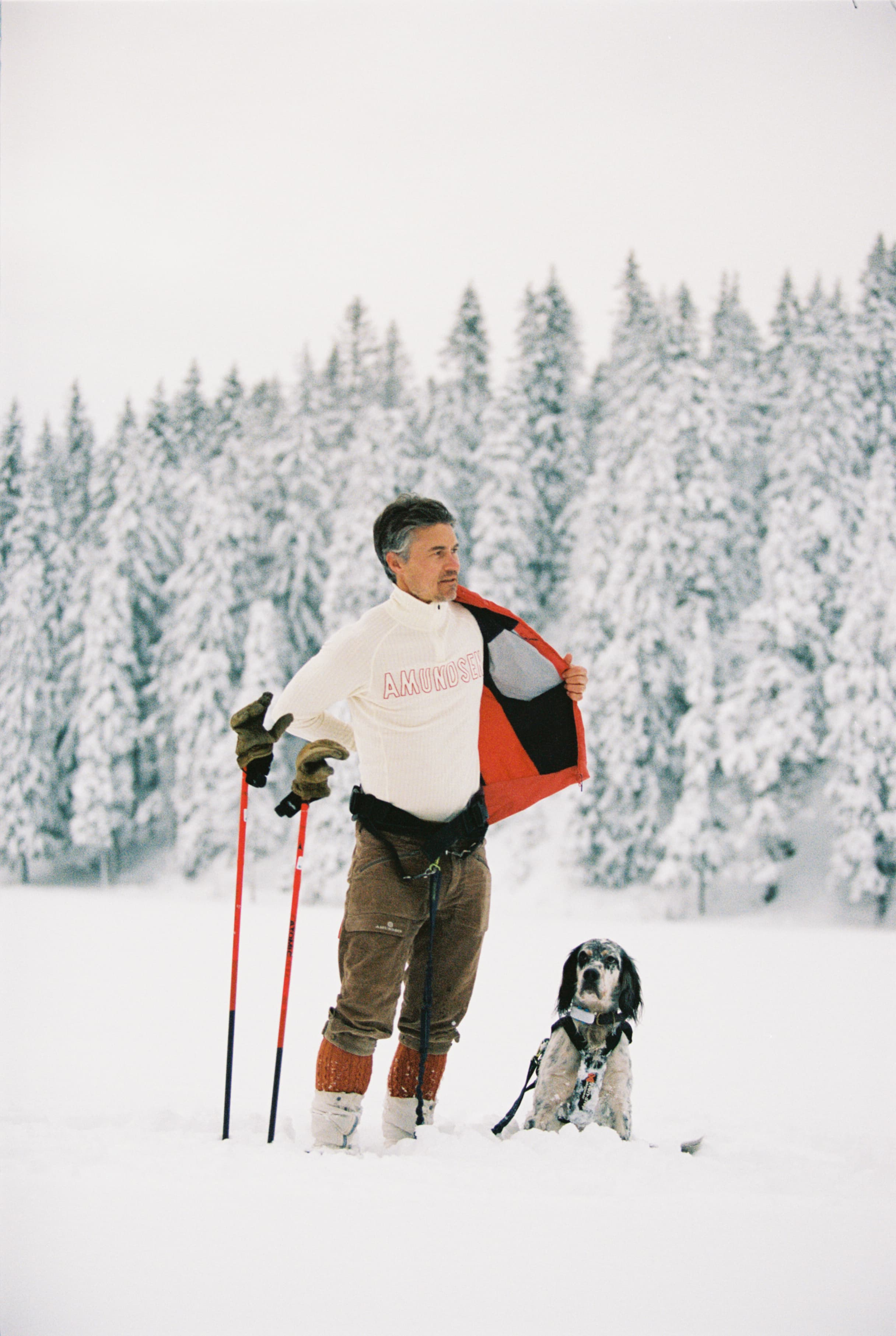 Man in winter attire stands with skis and poles, snowy forest background, dog sitting beside him.