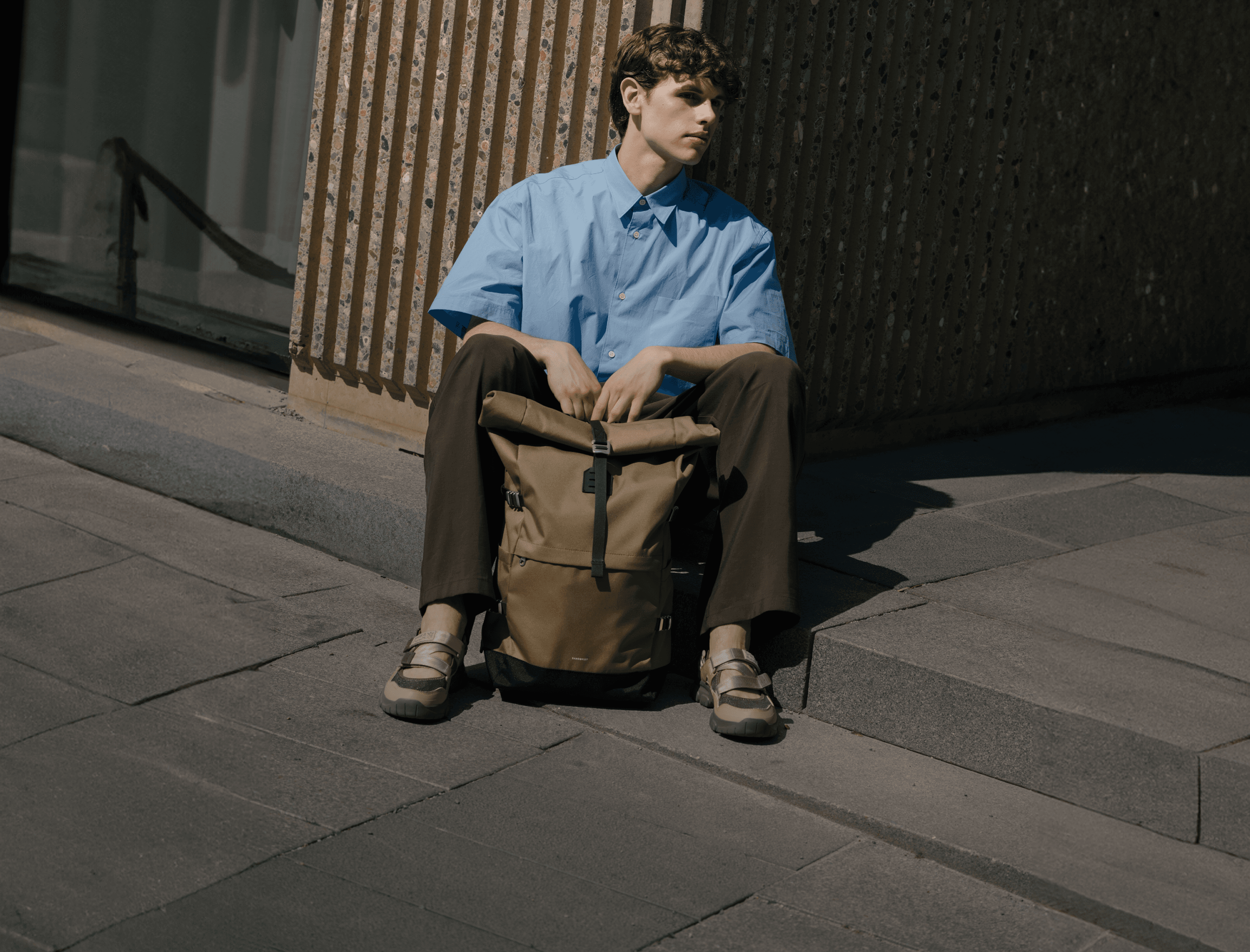 A person in a blue shirt and brown pants sits on steps, holding a tan backpack, with a modern building in the background.