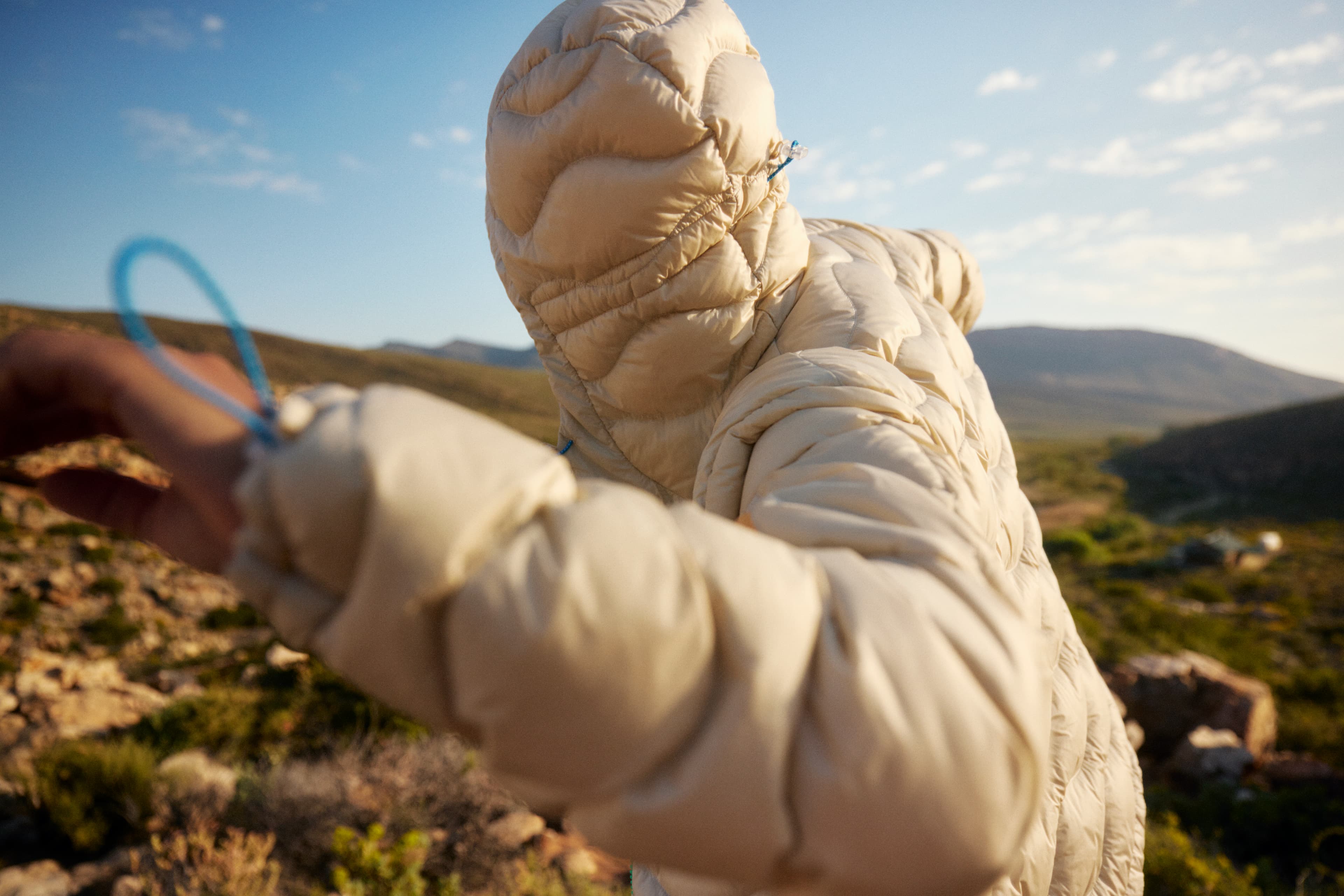 Person in a puffy beige jacket with hood up, standing outdoors in a rocky landscape under a blue sky.