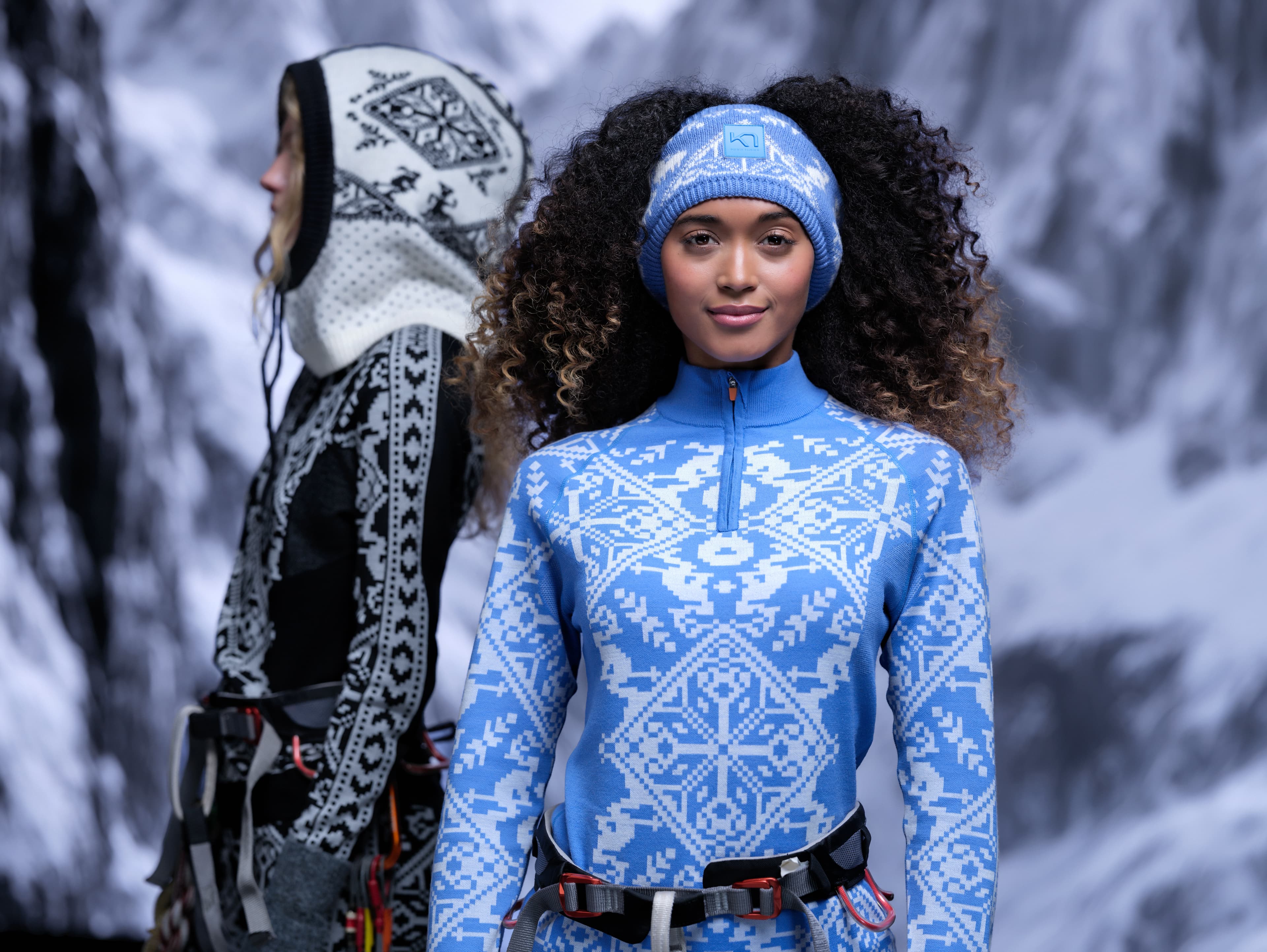 Two models in blue and black winter attire with intricate patterns, set against a snowy mountain backdrop.