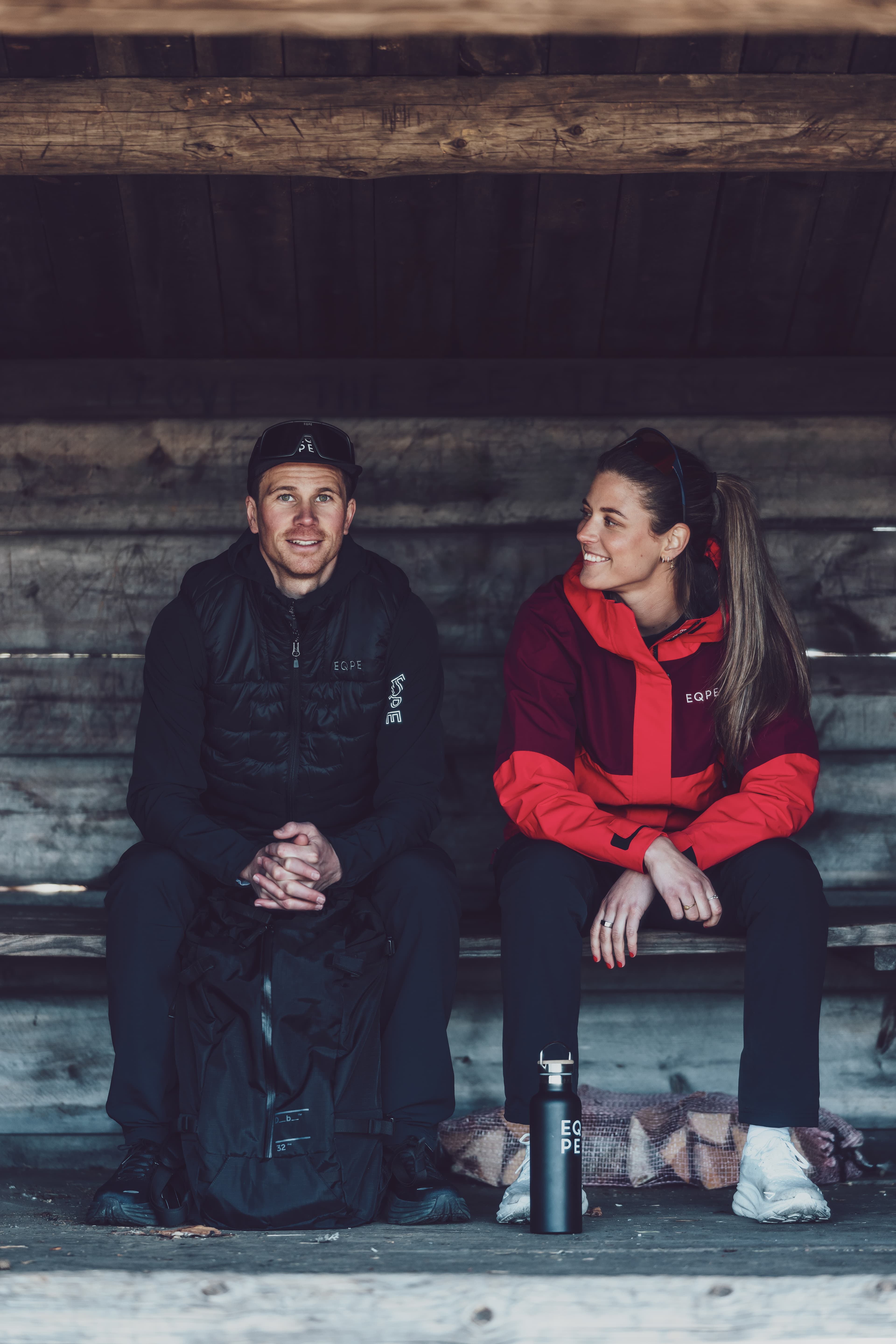 Two people sit on a wooden bench in a rustic shelter. The man wears a black jacket and cap, while the woman wears a red jacket, both smiling.