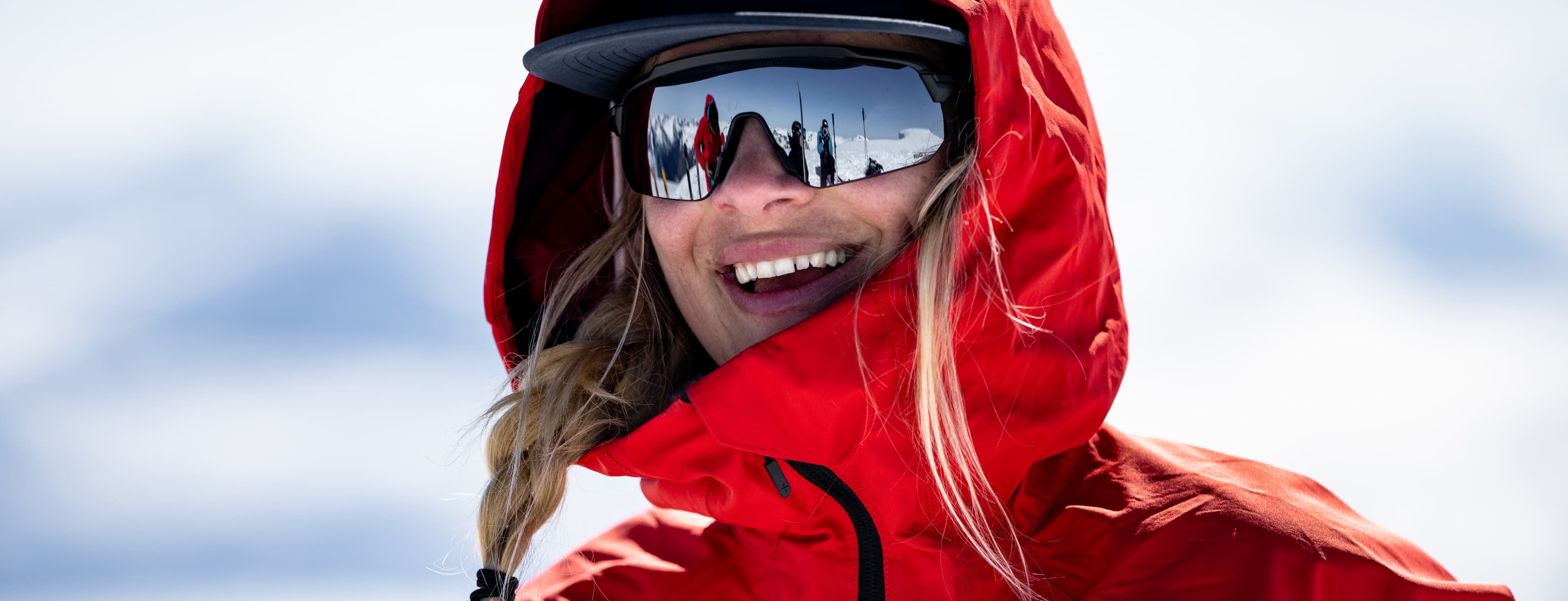 Person in a red jacket and sunglasses, smiling. Reflections of snowy scenery in the sunglasses.
