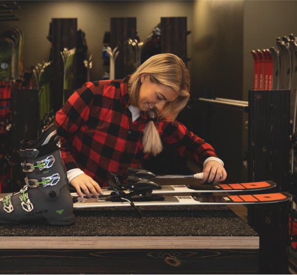 A person in a red plaid shirt adjusts ski bindings in a shop, with ski boots and skis visible on the counter.