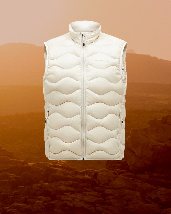 White puffer vest with wave pattern, sleeveless, front zipper, displayed against a warm-toned, rocky landscape background.