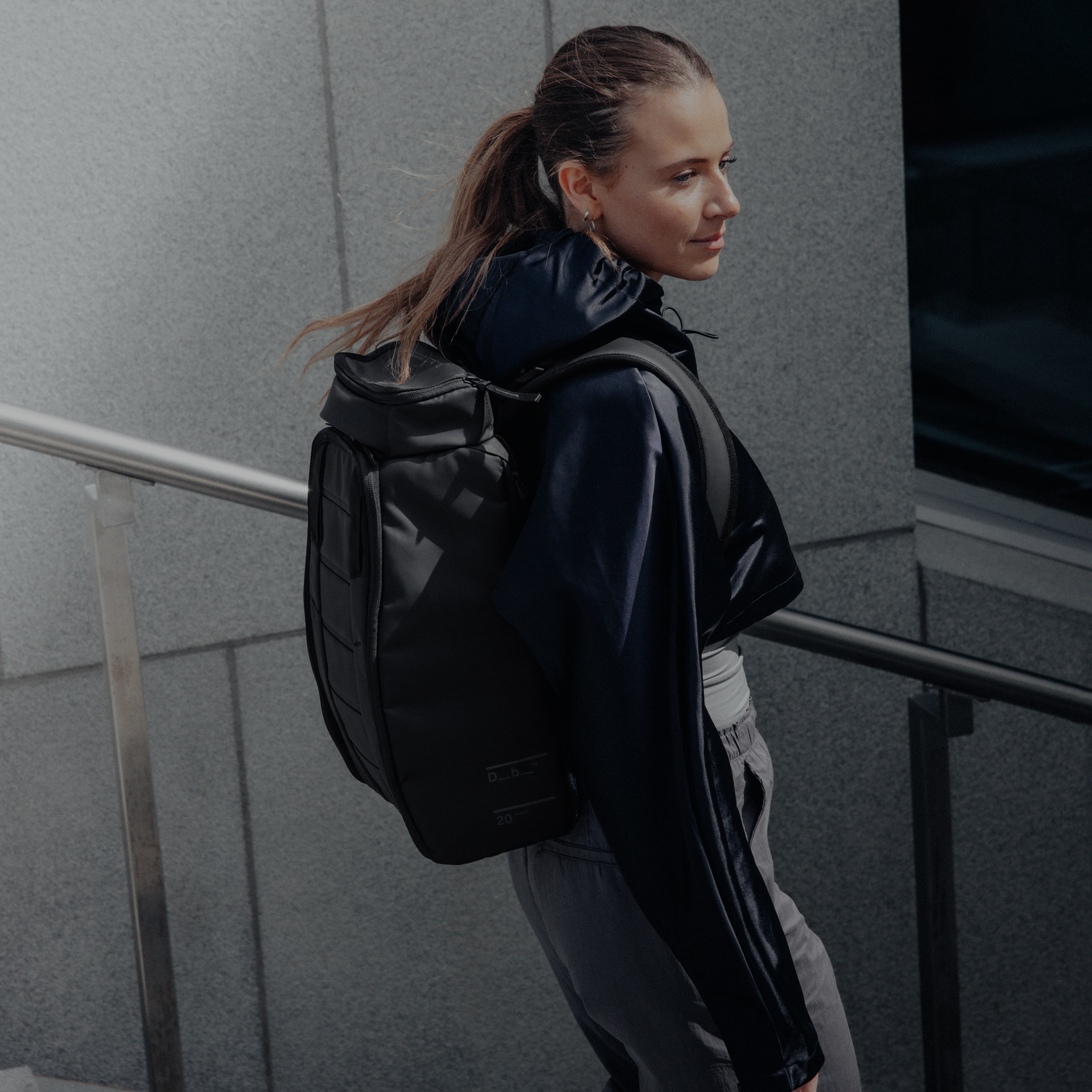 Woman with black backpack walks down stairs beside a gray building.