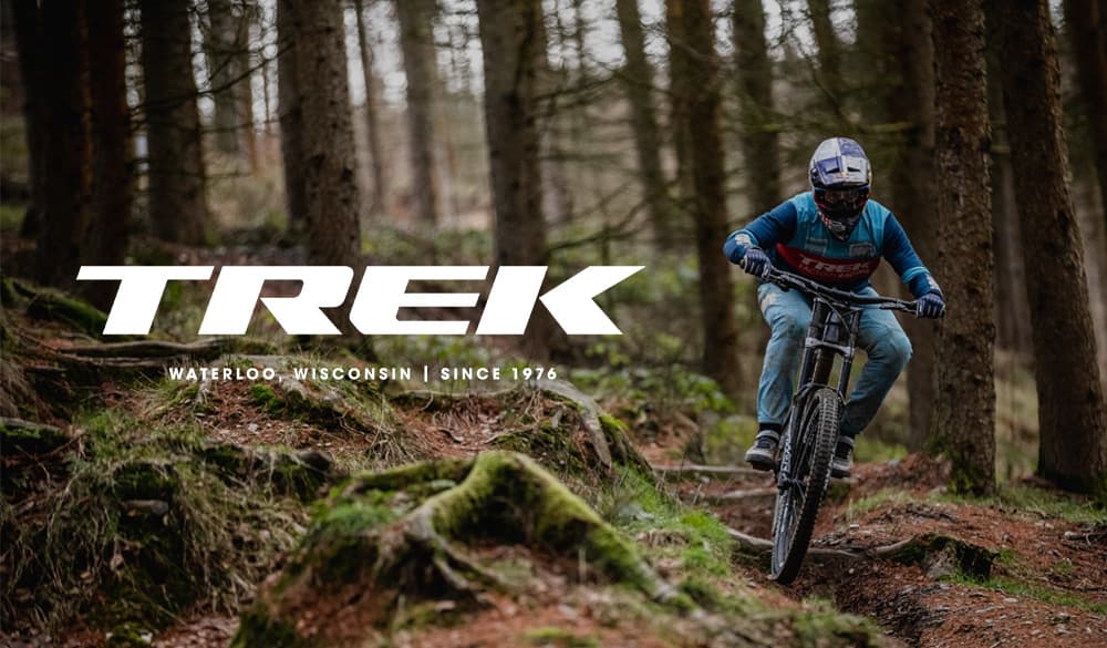 Mountain biker in a forest, wearing a helmet and blue gear, with "TREK" logo and location details in the background.