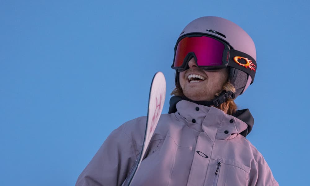 A person in a pink helmet and goggles smiles while holding a snowboard against a clear blue sky.