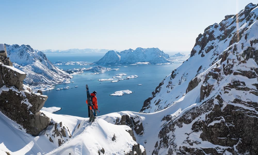 A skier in a red jacket stands on a snowy mountain ridge, overlooking a vast sea with distant snow-covered islands.