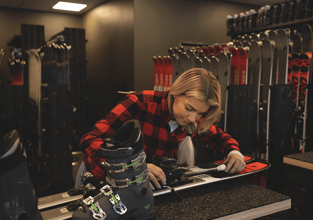 Woman in a plaid shirt adjusts ski bindings in a ski shop, surrounded by various skis and boots.