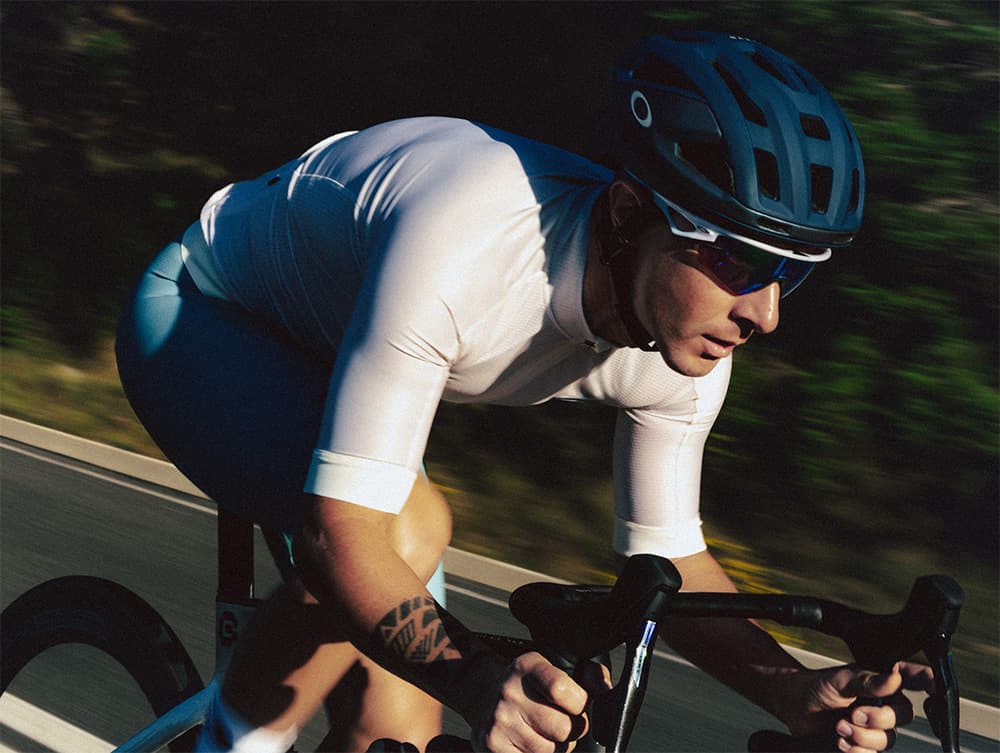 Cyclist in a white jersey and helmet rides a bike on a road, surrounded by blurred greenery, focused and aerodynamic.